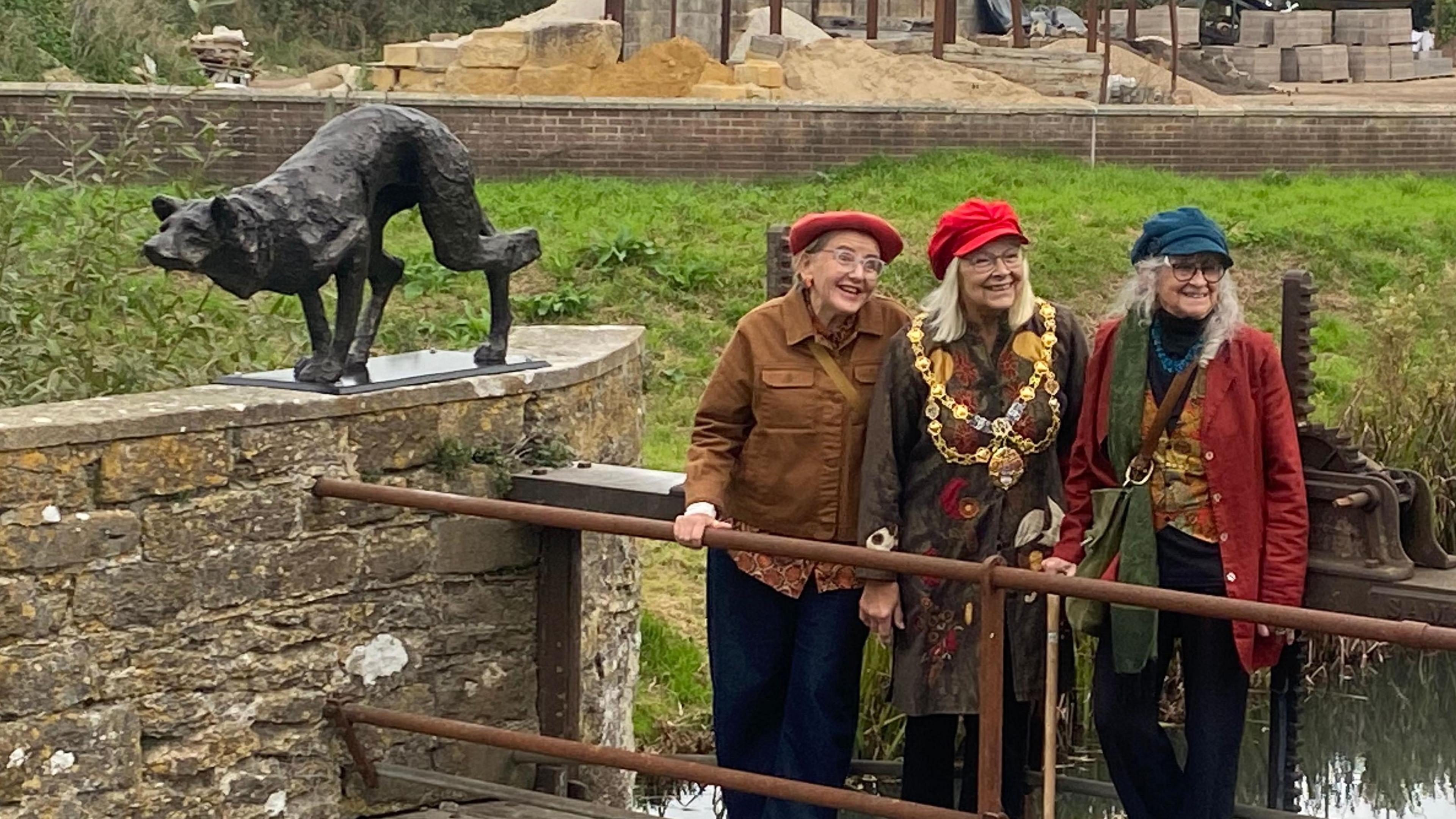 Ros Huxley, Anne Rickard and Greta Berlin pose for the cameras on the narrow wooden bridge over the weir next to the black sculpture of the stalking dog. Anne Rickard is wearing mayoral chains. All three are wearing glasses and soft peaked caps.