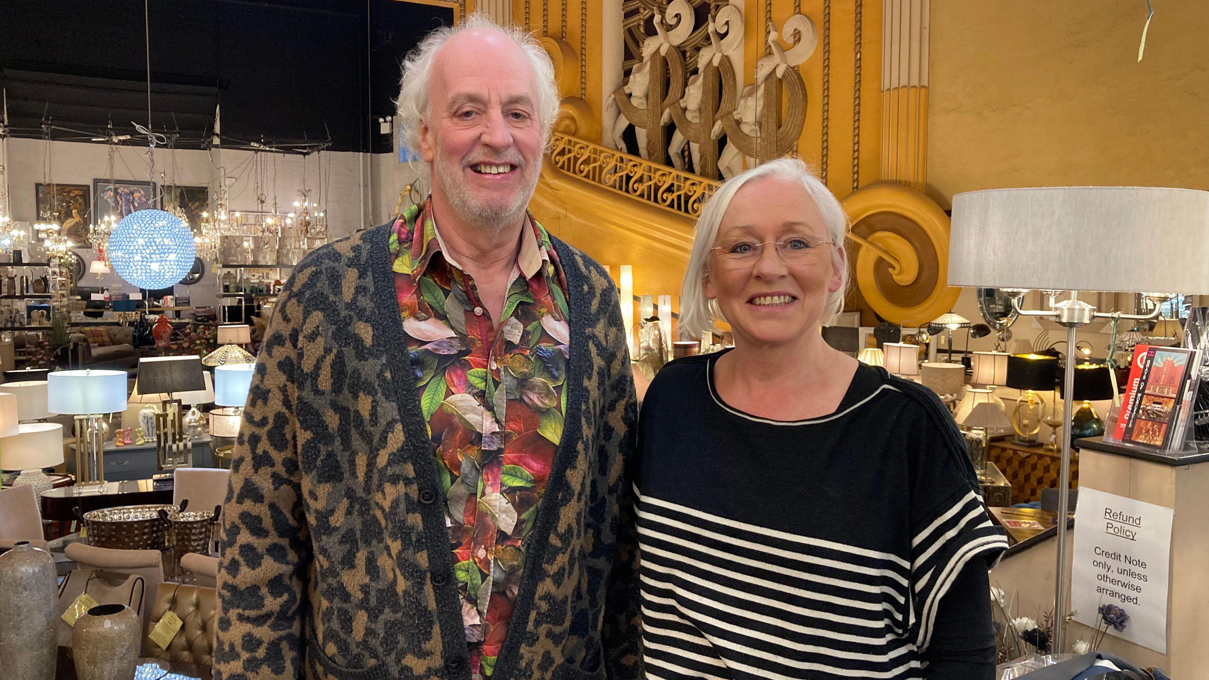 A man with white hair, wearing a brightly-colour shirt and cardigan, stands next to a white-haired woman wearing a black jumper. Behind them is an Art Deco sculpture, as well as furniture and several lamps.