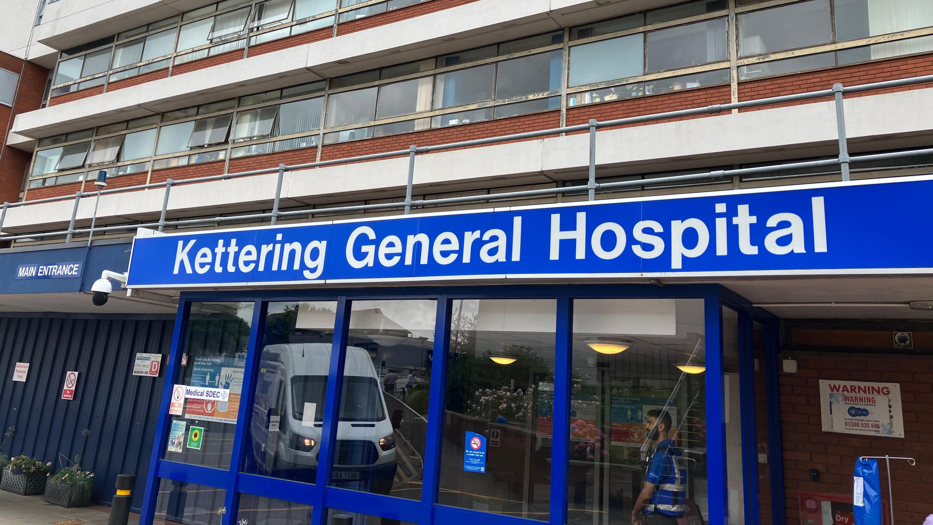 A blue entrance sign with Kettering General Hospital written on it with a security guard standing by the door. 