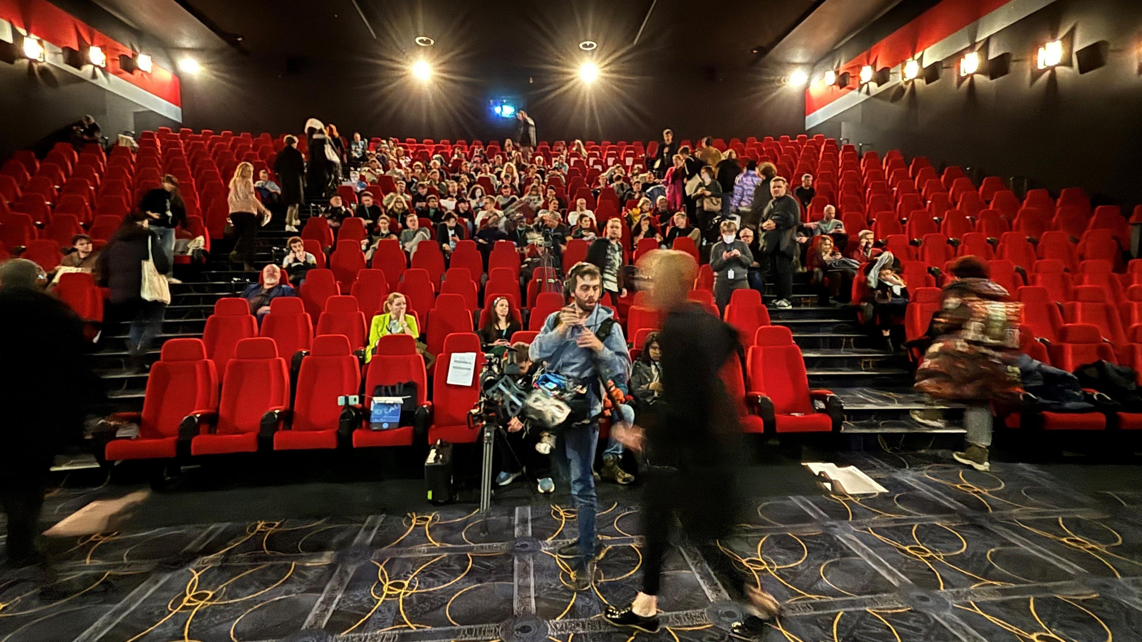 A cinema theatre with red seats and a patterned carpet. People take their seats.