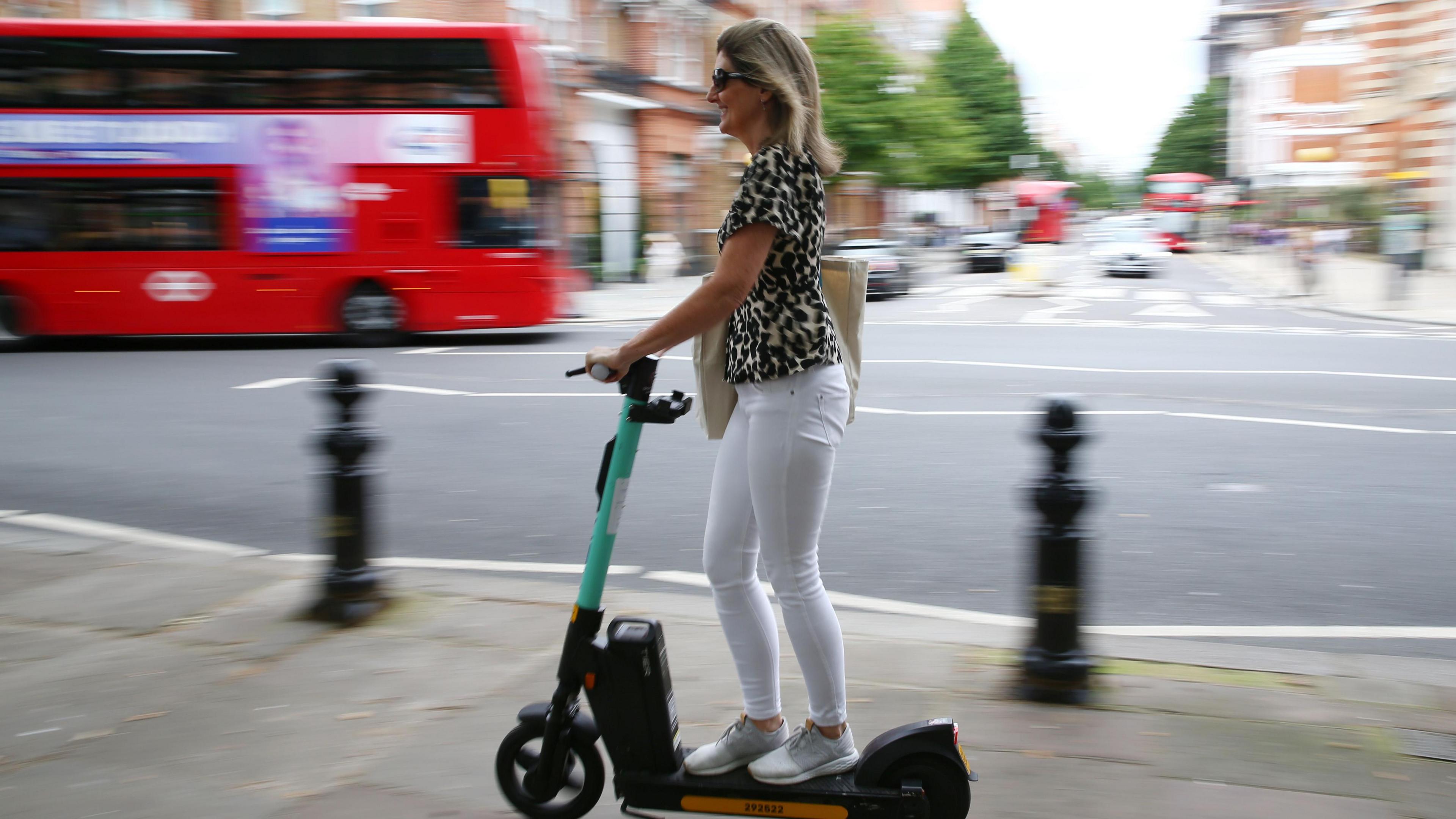 A blonde woman wearing white jeans, white trainers, a leopard-print top and sunglasses on a green and black e-scooter 