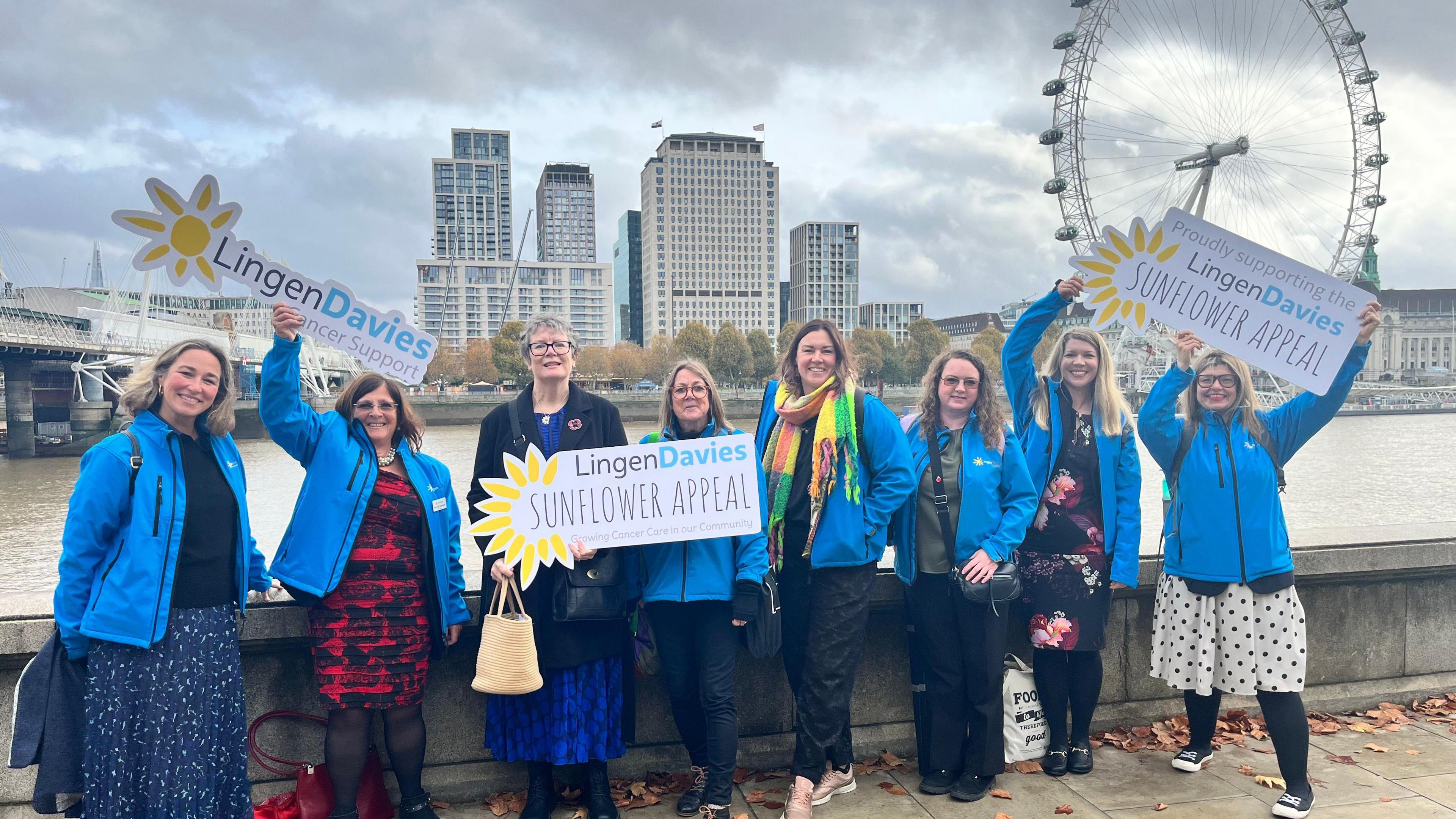 Eight women wearing blue jackets and holding signs reading "Lingen Davies Sunflower Appeal" stand in front of the London eye and high-rise buildings with the River Thames behind them.