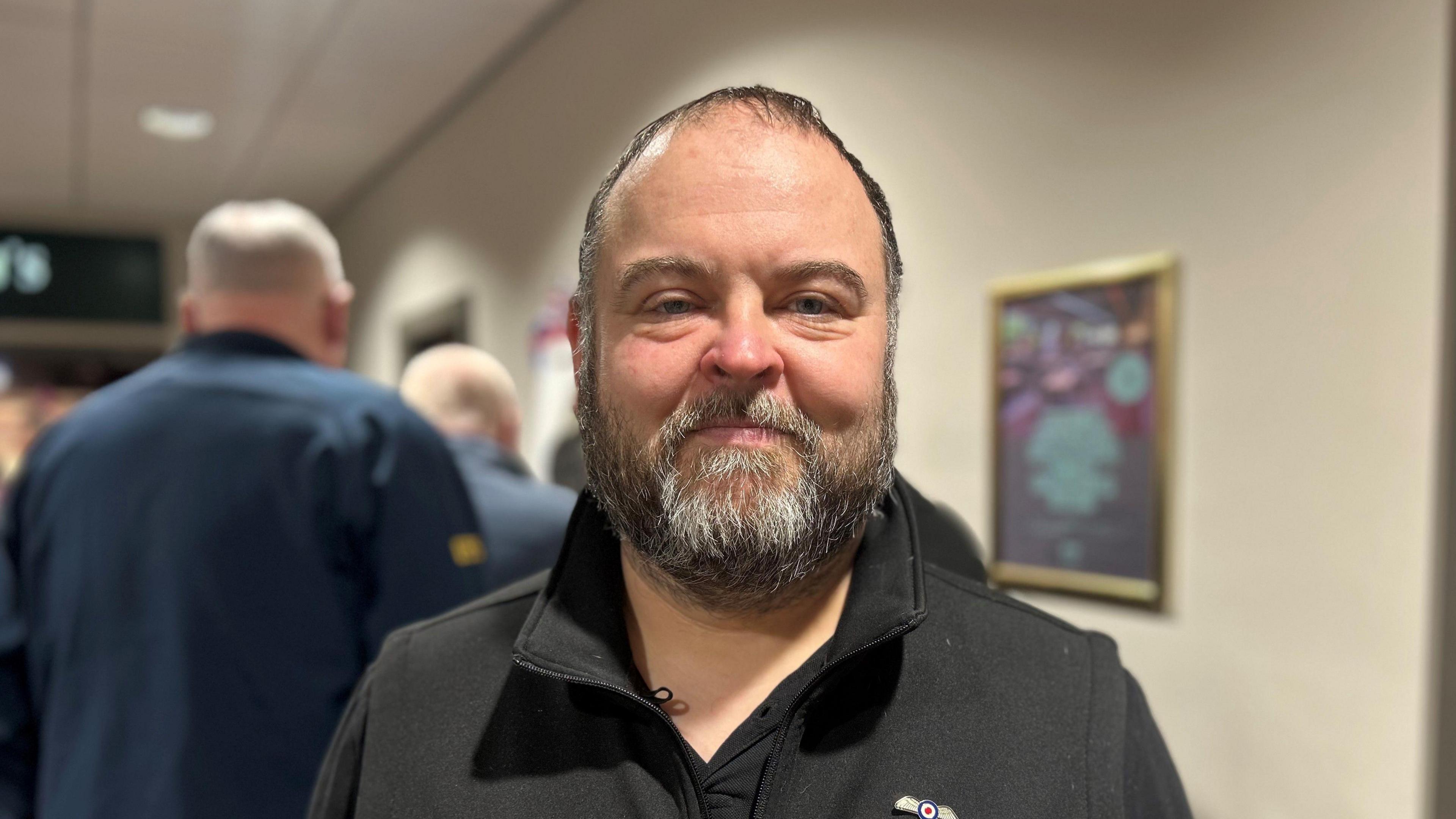 David Bygate-Pittiglio has a beard which is slightly graying. He is wearing a black top with an RAF symbol on the right hand side. It has an open collar and his hair is short. 