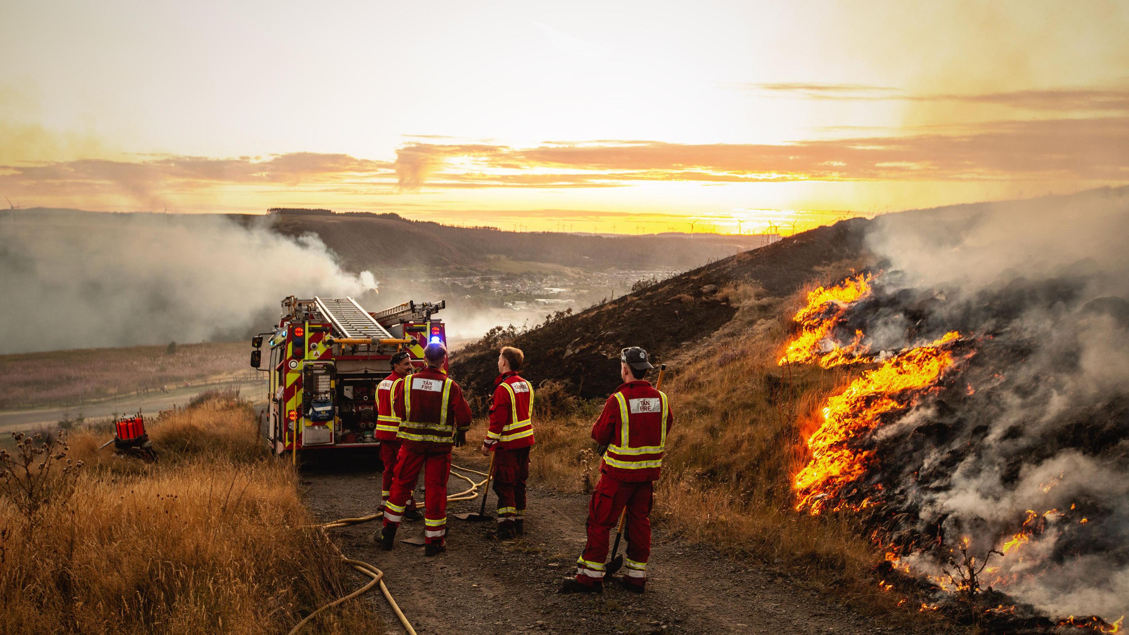 Firefighters are stood next to very dry grass, which some if it has have been burnt or is on fire. A fire truck is pictured on a path, and the countryside in the distance has smoke coming from the ground.