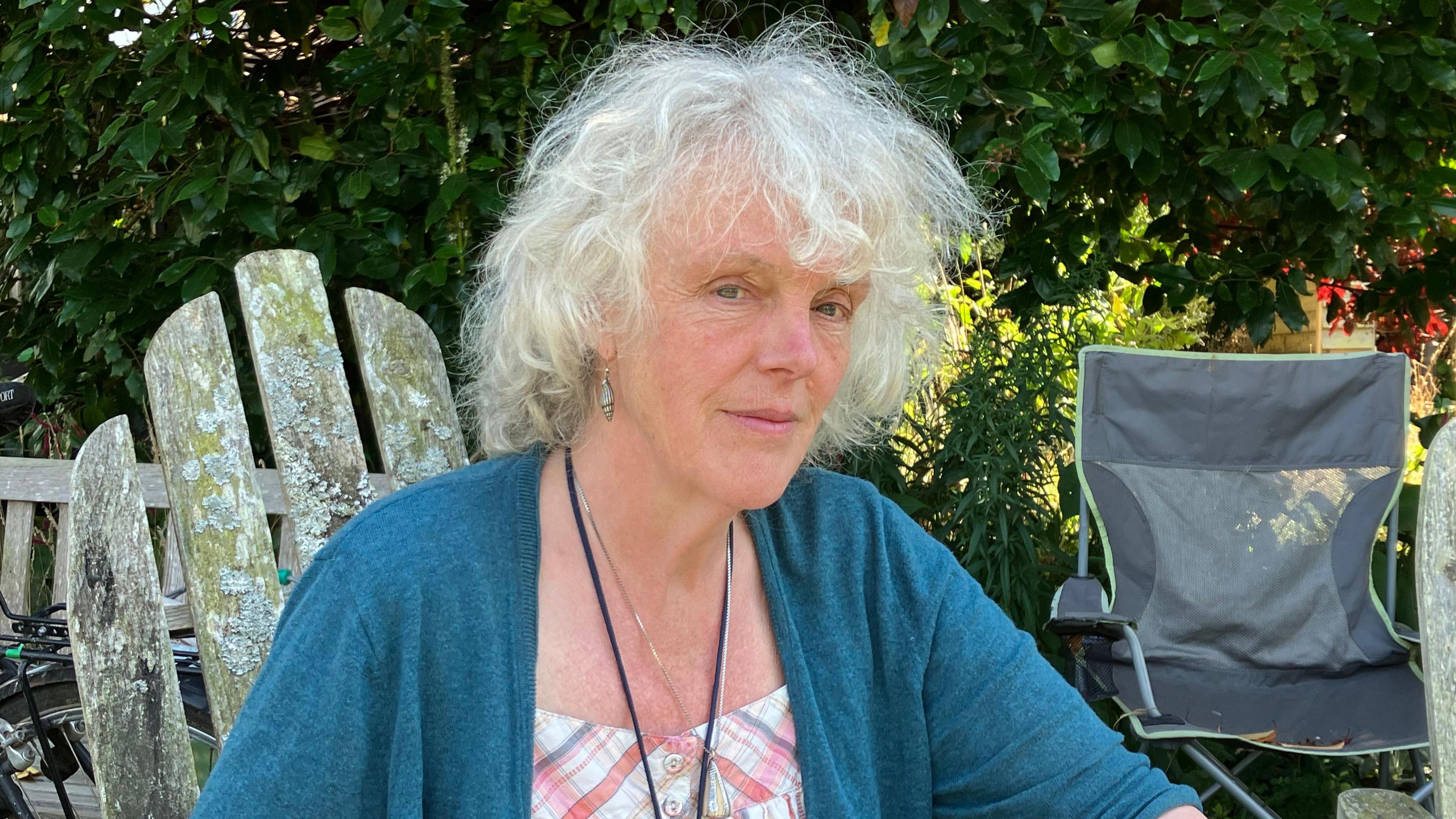 A village resident with white shoulder length, wavy hair, wearing a blue cardigan and earrings sits in a wooden chair in a sunlit garden