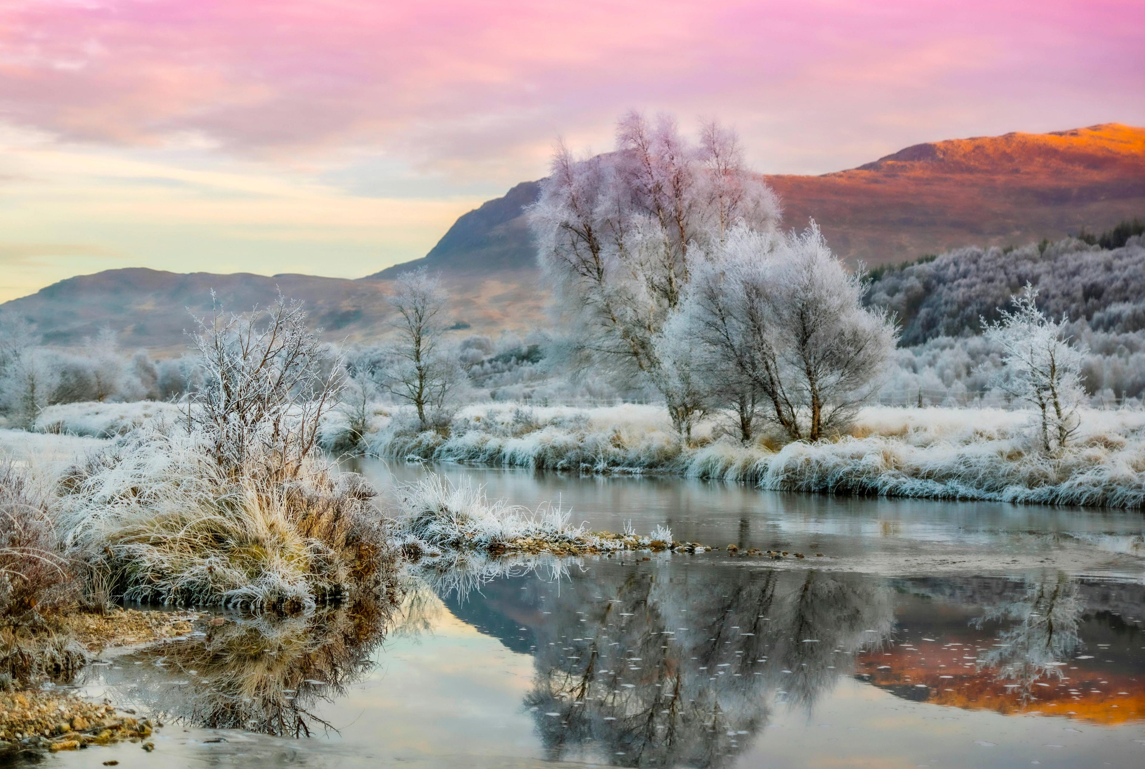 Landscape image of a pale pink sky over the hills with frost-covered trees and grass below next to a river