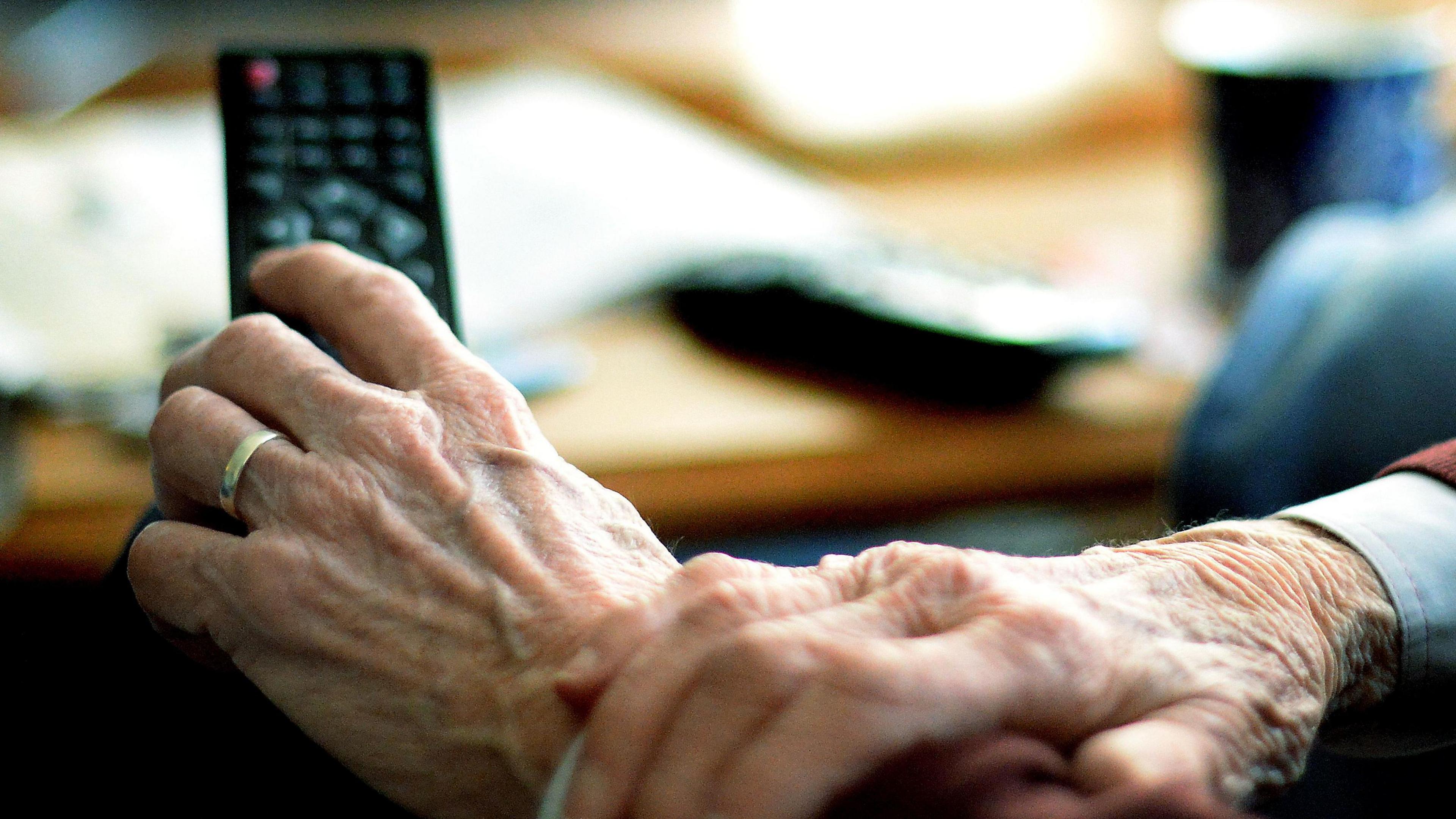 Close-up of the hands of an elderly person holding a television remote control. In the background there is a coffee table, upon which there is a coffee table, papers and other items.