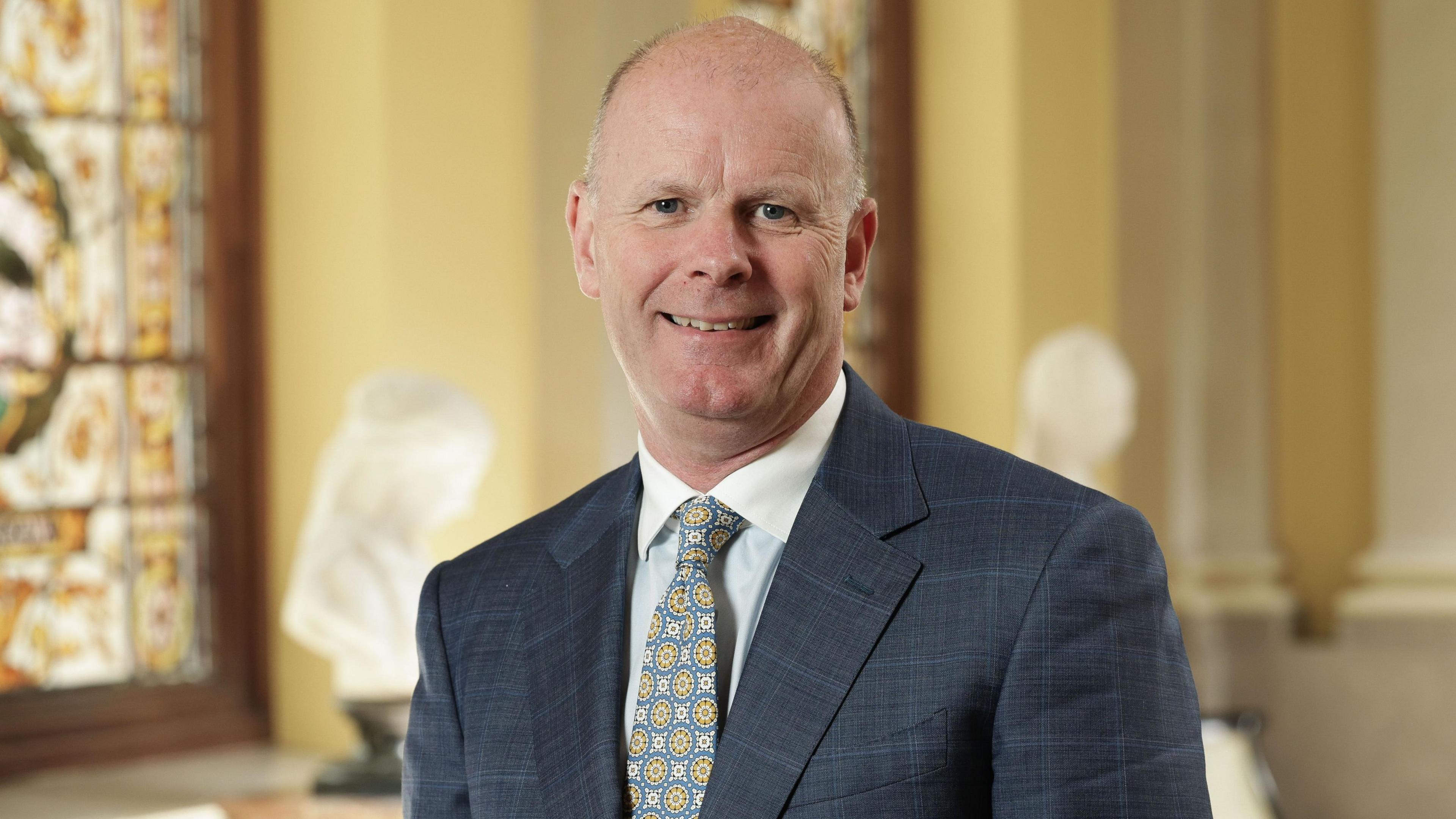 A man in a blue suit with a blue and yellow patterned tie. He is wearing a white shirt.