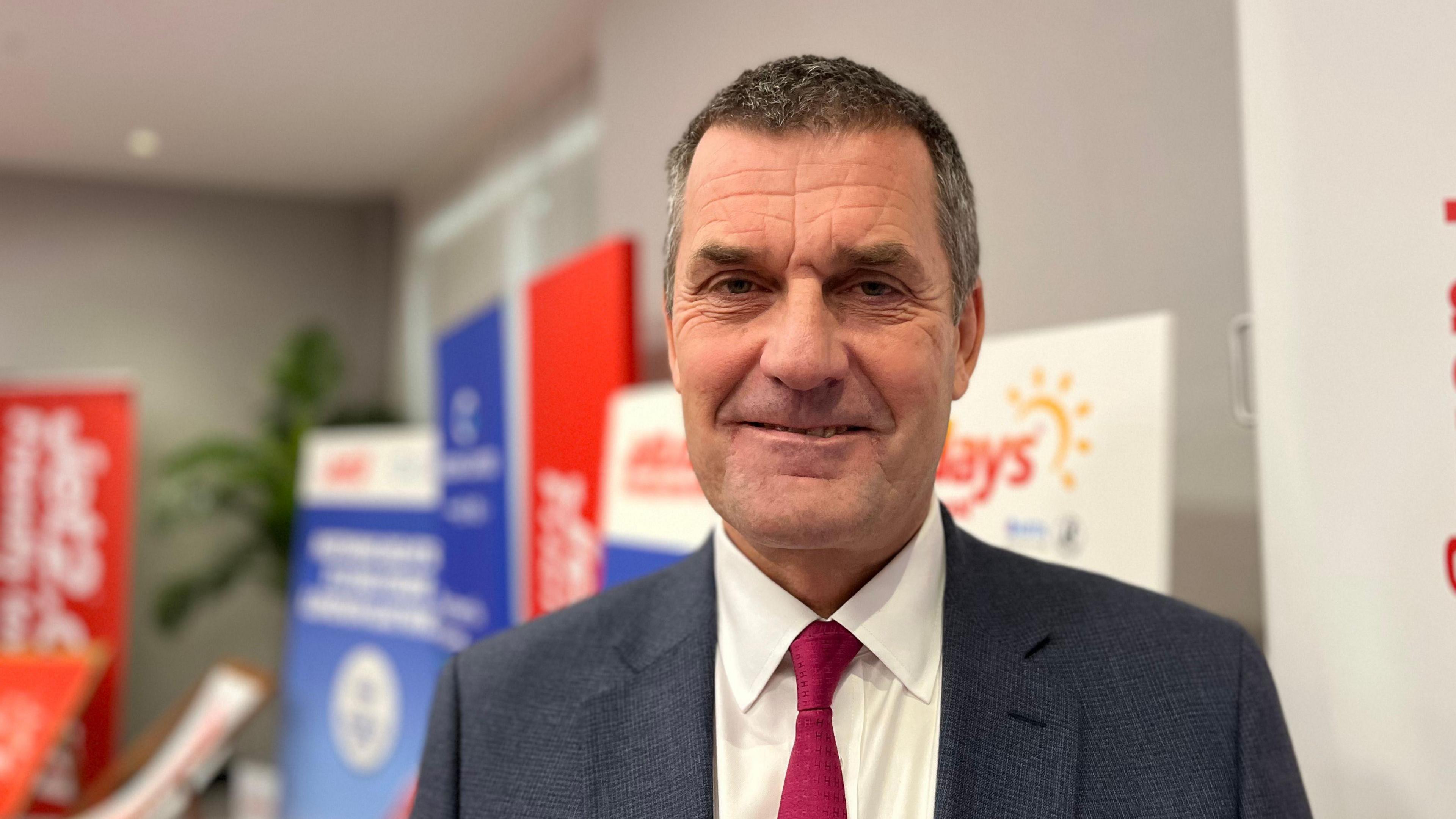 A man with short dark hair, greying at the temples, in a navy suit, with a white shirt and red tie.