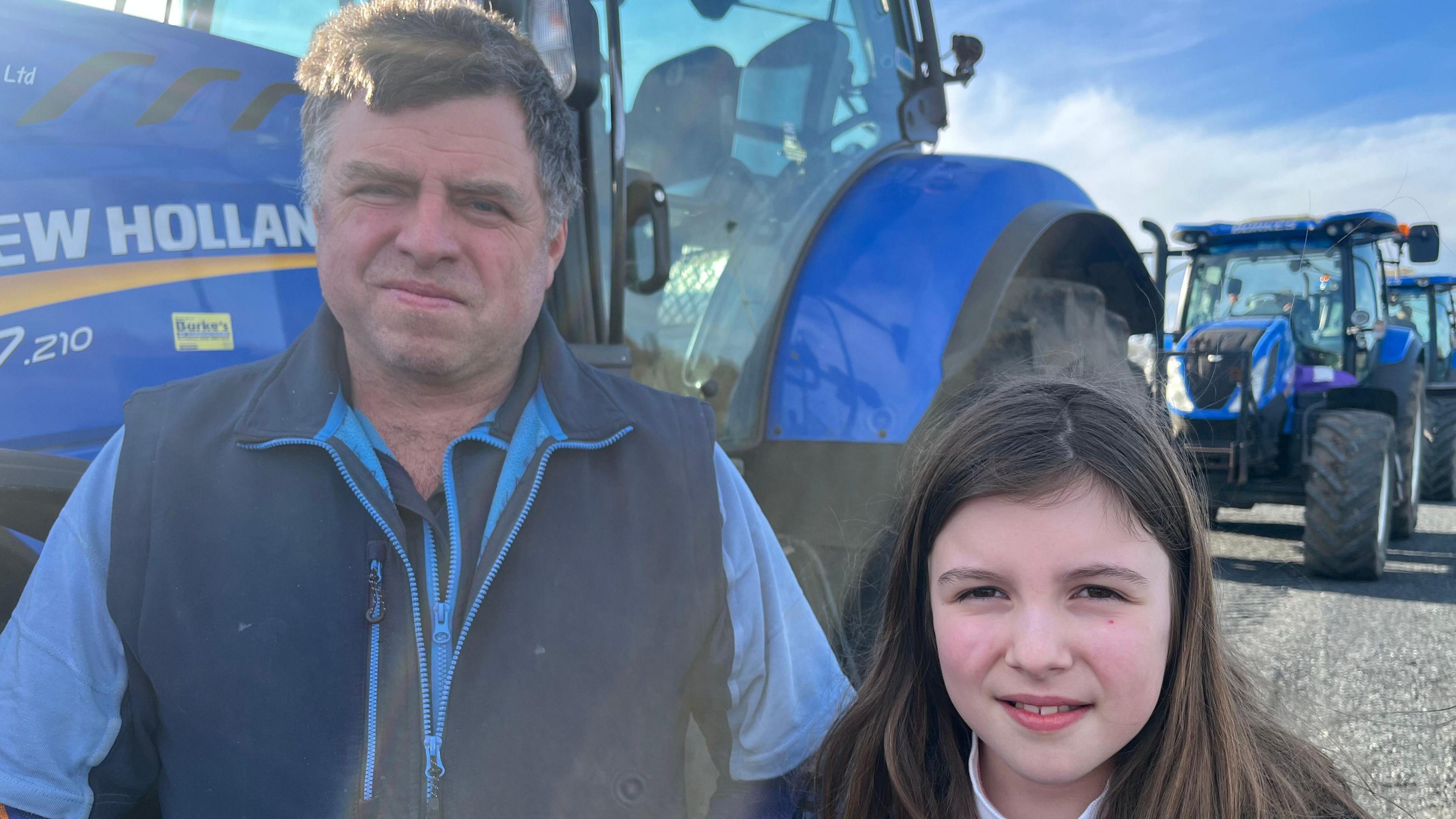 Carl, left, is standing in front of a blue tractor. He is wearing a navy fleece body warmer and light blue short sleeve top. Annabelle, right, has long brown hair and looking directly at the camera. 