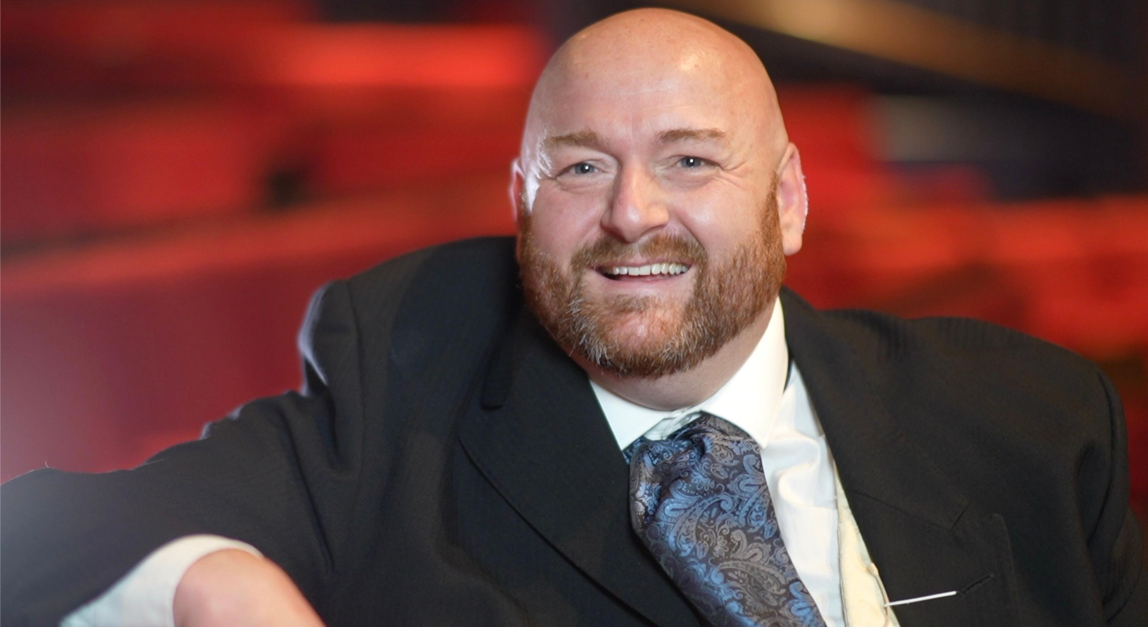 Mr Fosker is pictured smiling at the camera while sitting in The Seckford Theatre, in Woodbridge. He is bald and has a light brown beard. He is wearing a black suit and a white shirt.