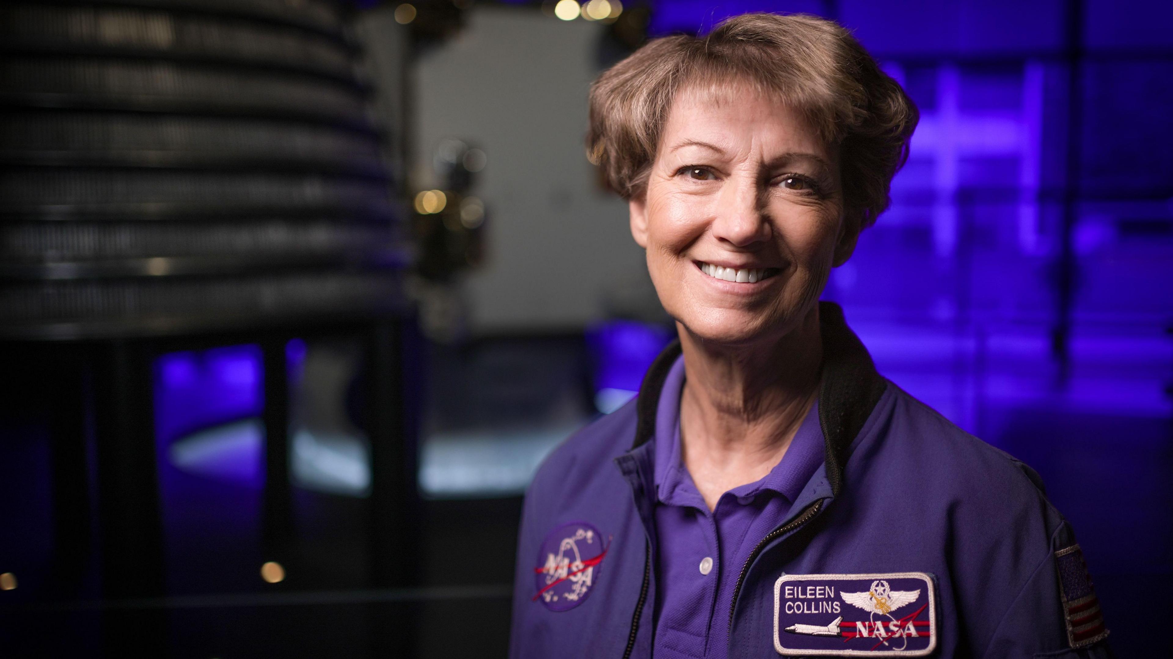 Eileen Collins wearing a blue jacket with a round Nasa patch on one side and a rectangular shuttle patch on the other with her name. She has short brown hair and brown eyes and is smiling as she looks directly to camera. She is in the space gallery at the Science Museum and an out of focus rocket engine can be seen behind her.