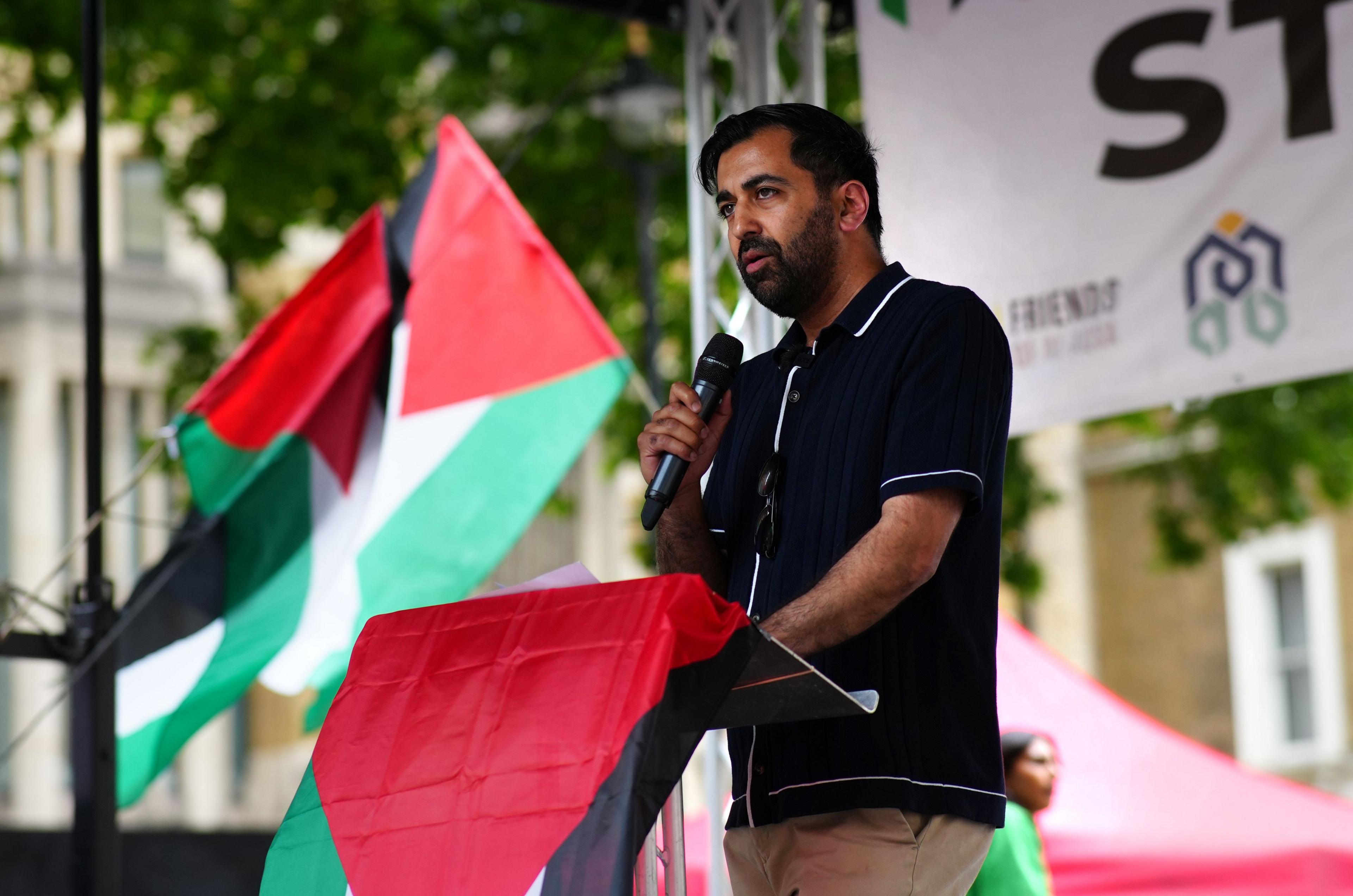 Humza Yousaf, who has dark hair and a dark beard, is standing on a podium while speaking into a microphone. He is dressed in a black polo shirt and sand-coloured trousers. There is a red, white, green and black Palestine flag draped over the podium and two more flying in the background