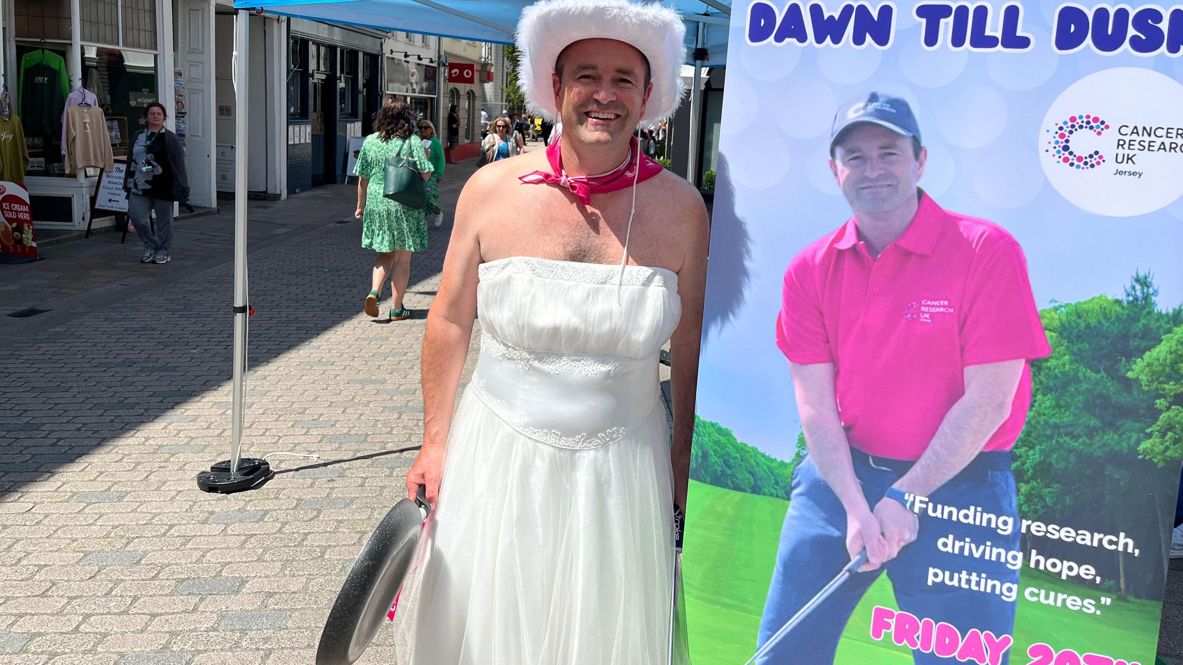 A middle-aged man in a white wedding dress, with a fluffy white cowboy hat and a pink neckerchief stands next to a roller banner with a picture of himself playing golf in a pink polo shirt. Text reads: Olly's Golf Challenge Dawn Till Dusk. Cancer Research UK.