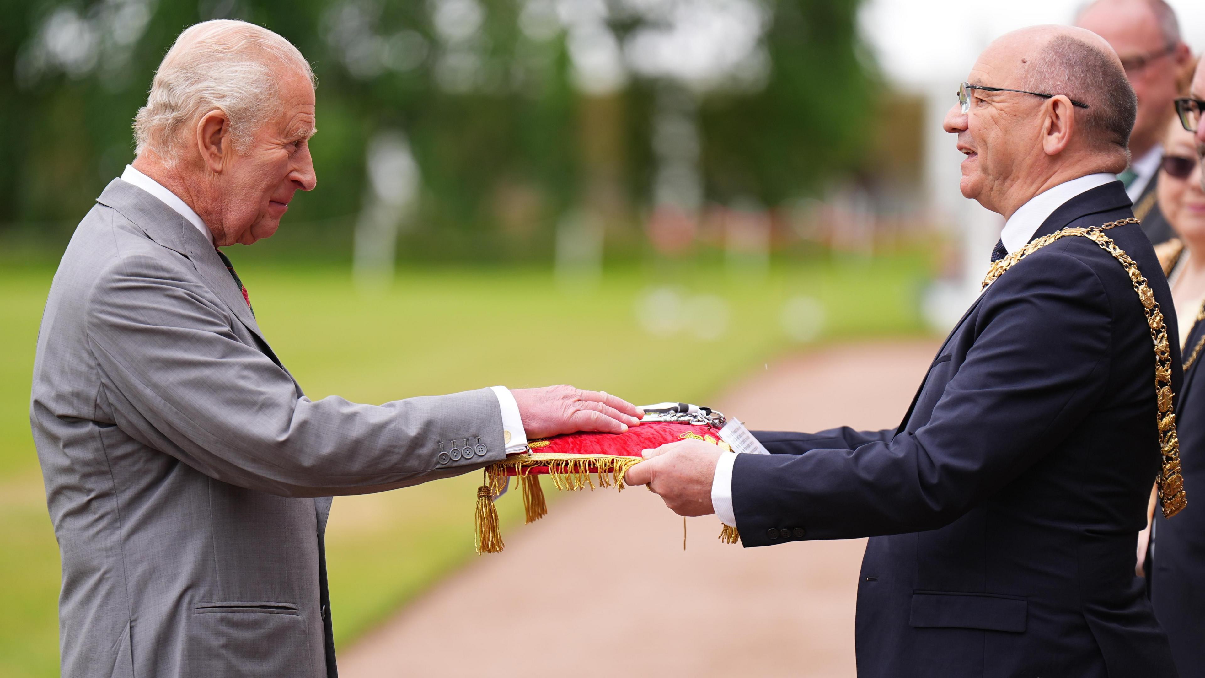 King Charles III wearing a grey suit and white shirt places his hand on a red cushion held by the Lord Provost of Edinburgh Robert Aldridge, who wears glasses and a dark-coloured suit. 