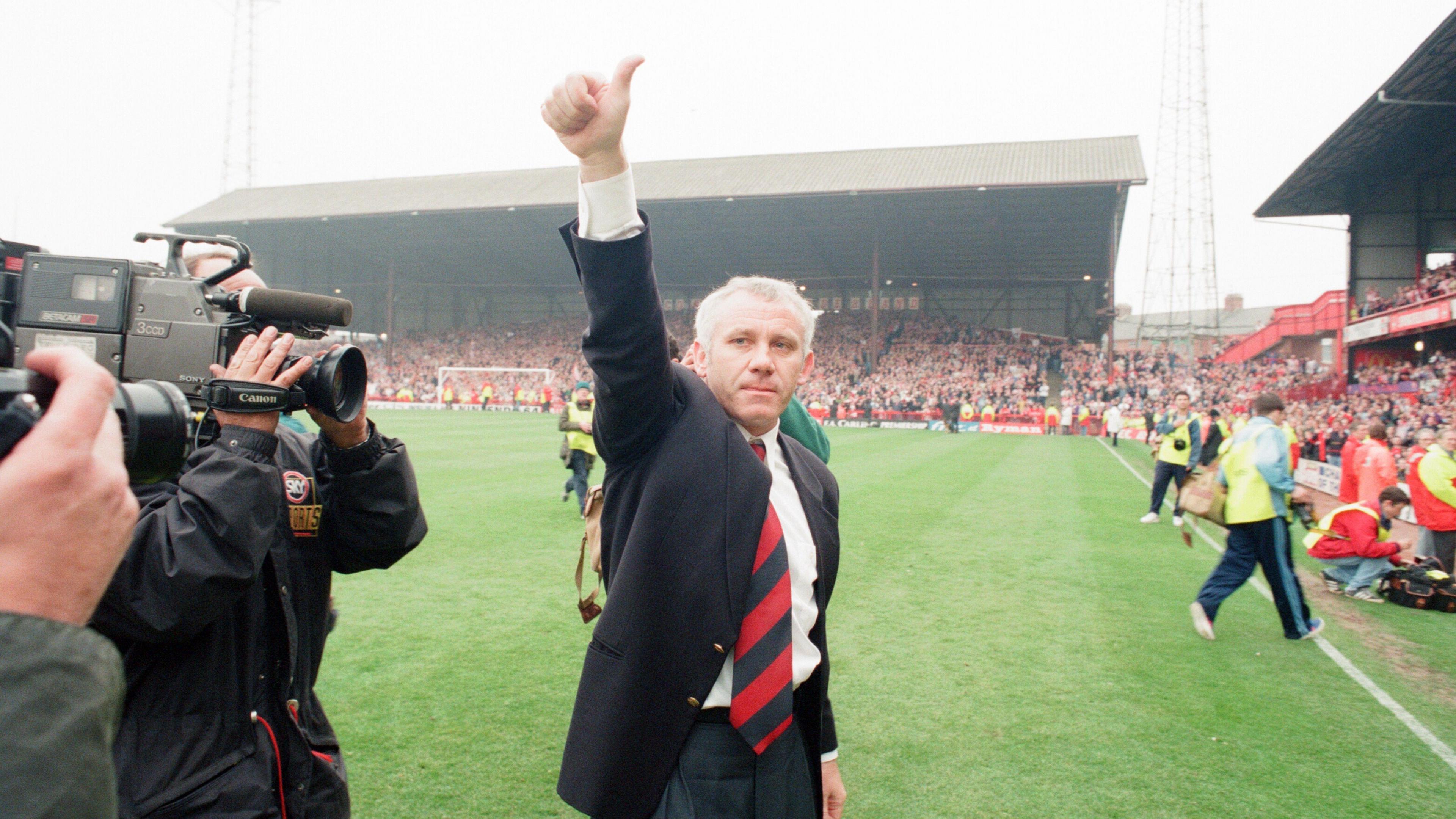 Peter Reid gives a thumbs-up gesture as he leaves the pitch at Roker Park following Sunderland's final game at the stadium. He has short grey hair and is wearing a dark blazer, white shirt and red tie. Two television cameramen are filming him. Thousands of fans are in the ground's stands.