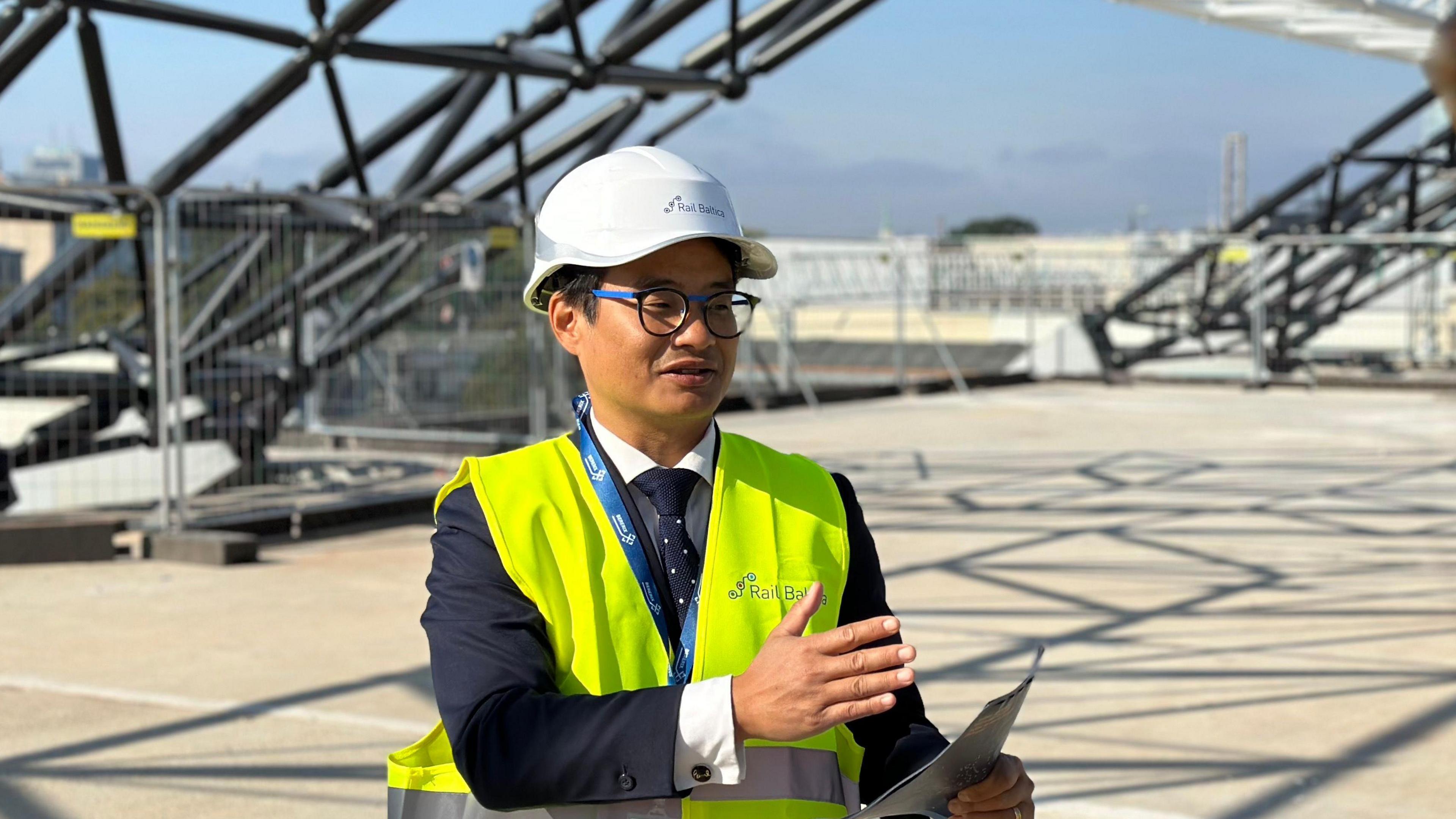 French engineer Emilien Dang, in a neon vest and white hard hat, gestures with a piece of paper in his hand. He shows the BBC the new railway terminal being built in Riga, with metal structures rising behind him.