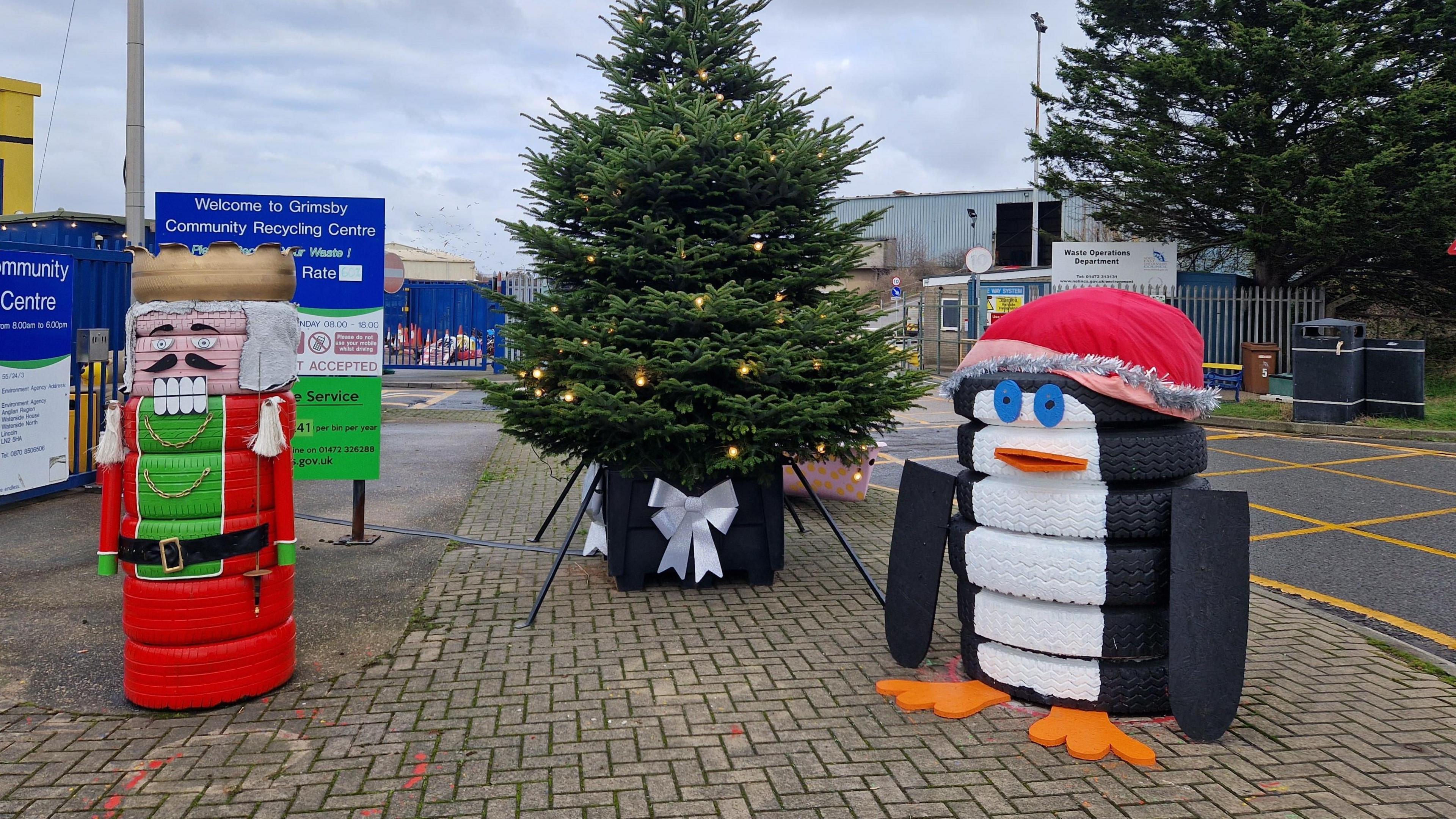 Two stacks of recycled tyres stand either side of a large Christmas tree decorated with lights. The eight tyres on the left side of the image have been stacked up and painted in gold, red, green and pink to resemble a traditional nutcracker doll. The seven tyres on the right side of the image have been made to look like a penguin wearing a red hat.