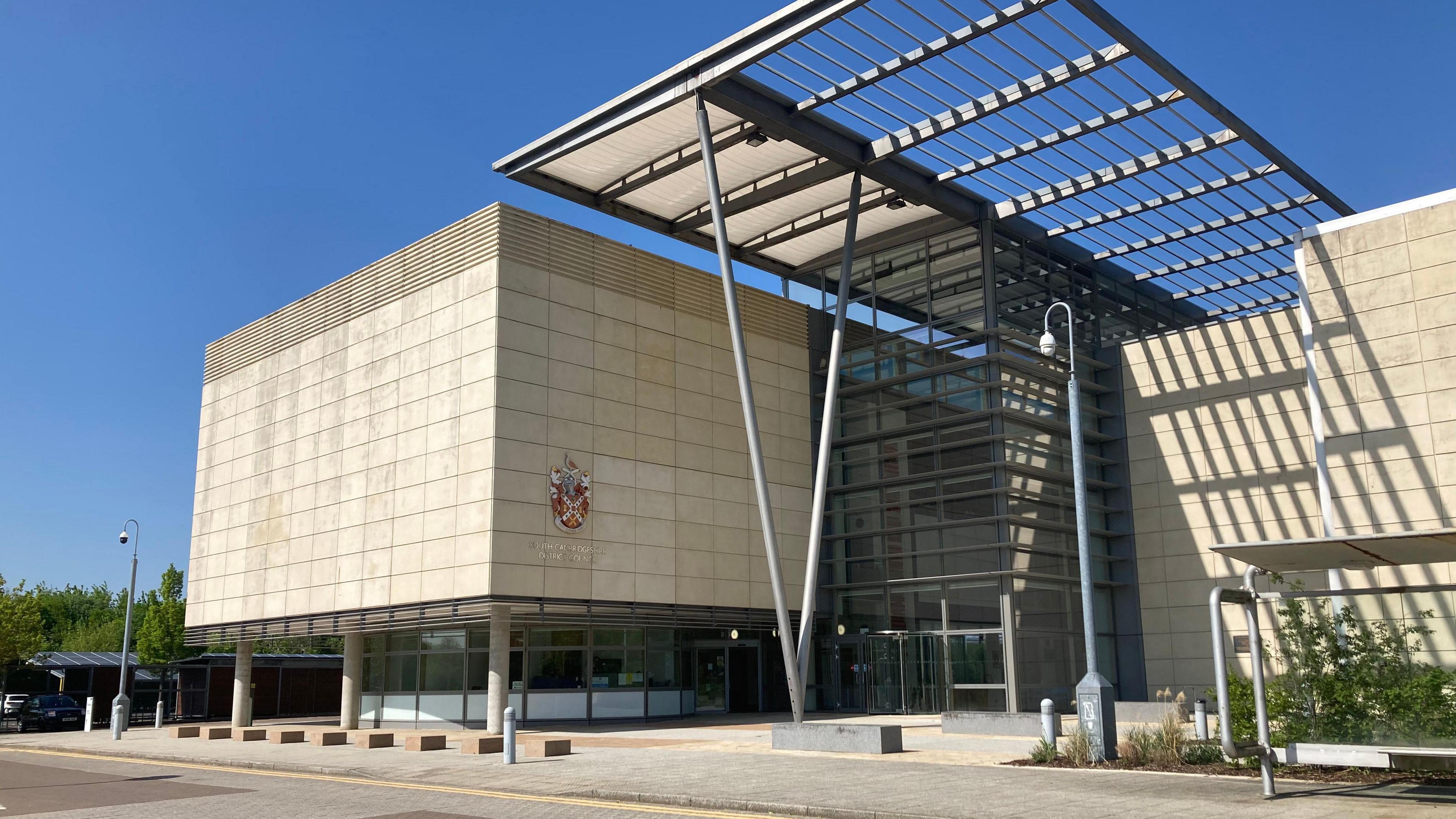 A large beige building with a council coat of arms and the words South Cambridgeshire District Council written on it. A glass fronted entrance is next to it with a further beige building attached to the other side. A bright blue sky is behind the building.