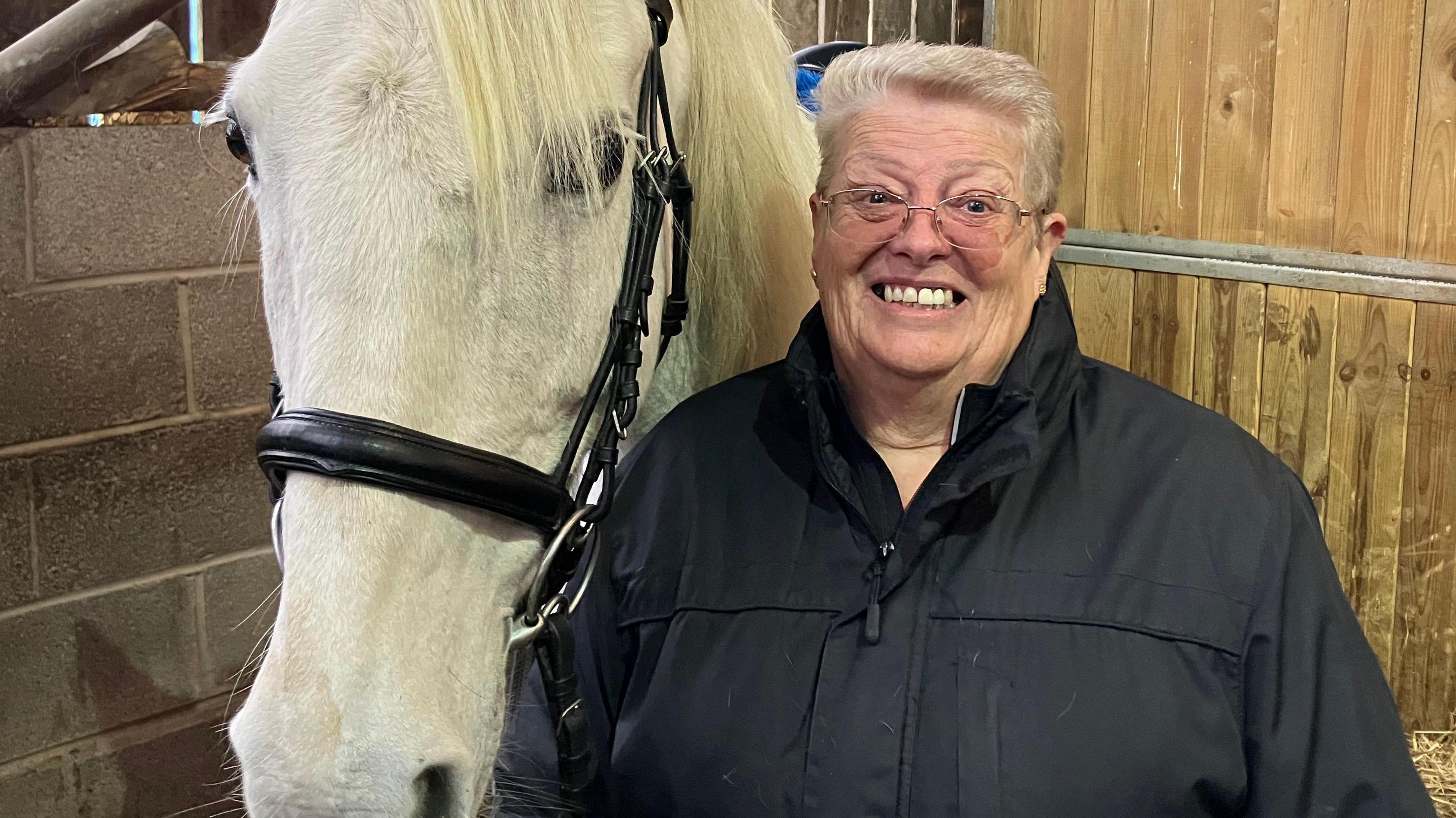 Wendy stands next to Seb the horse at the stables. Wendy is smiling wearing a black rancoat, and Seb's white head is visible next to her.