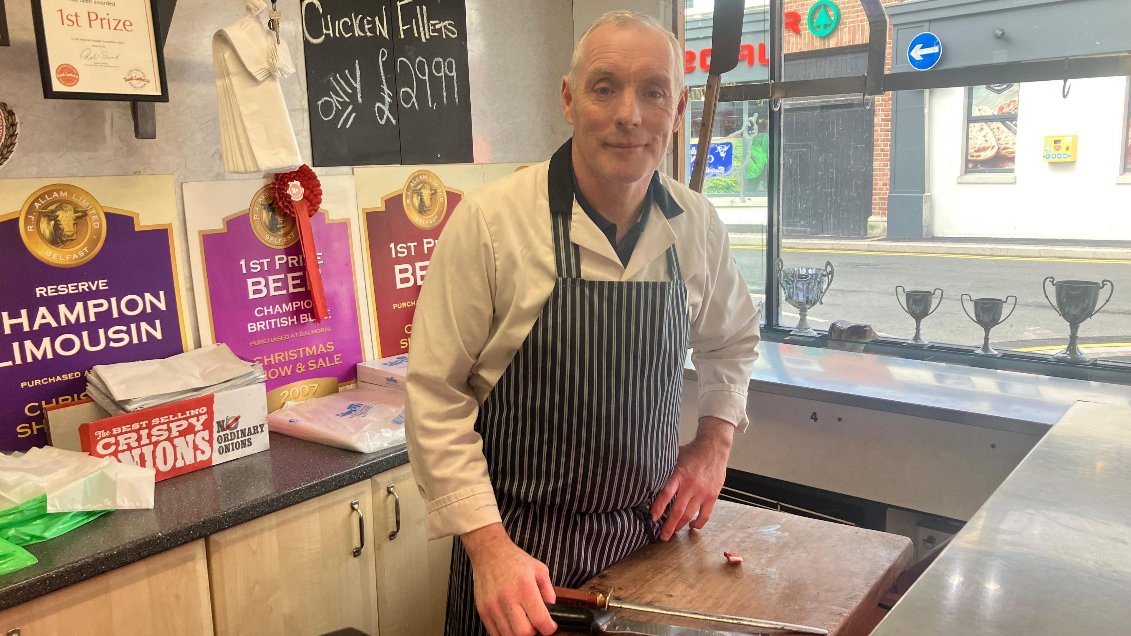 A man stands in a butcher shop wearing a white shirt and a black and white striped apron 