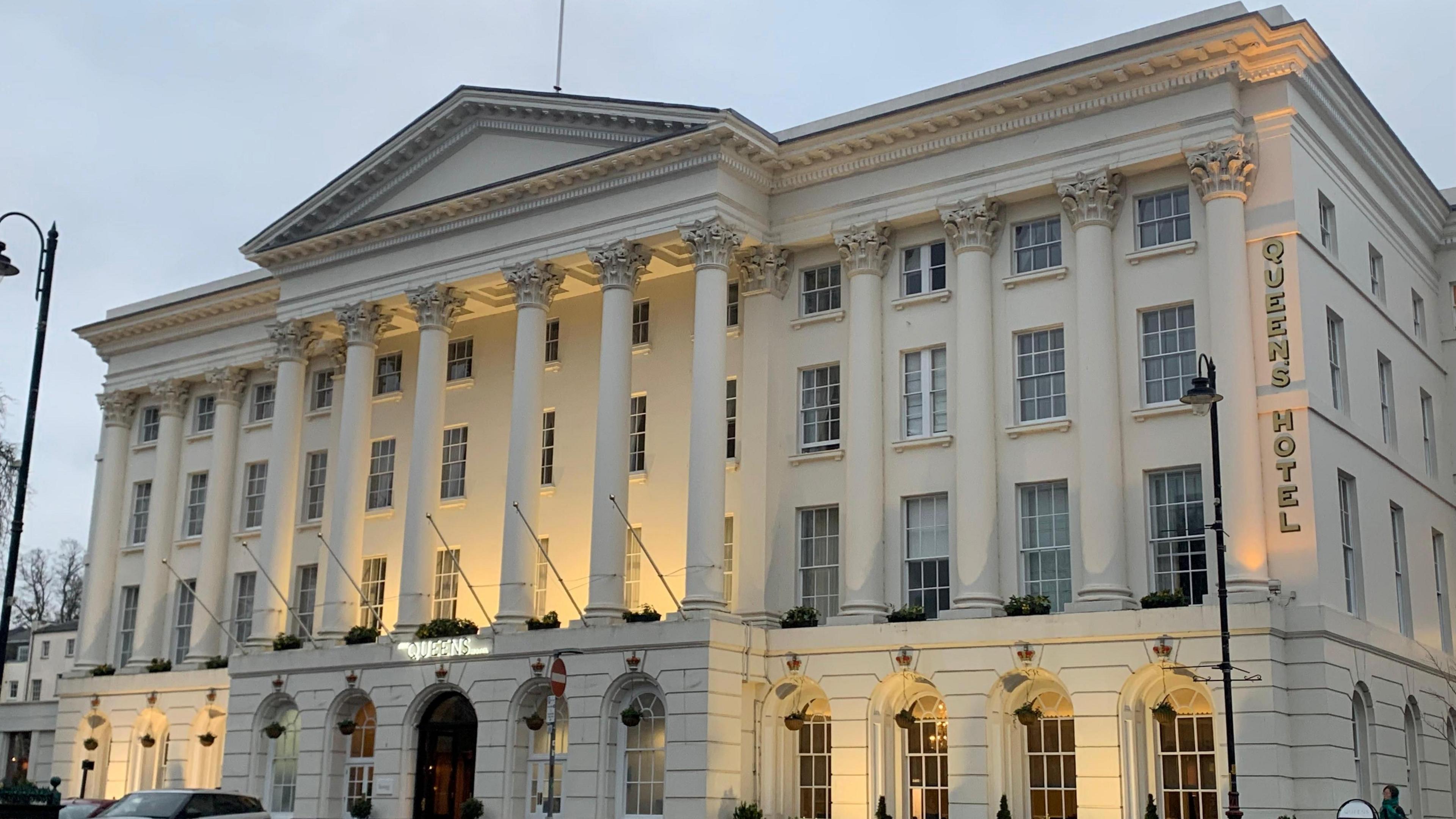 The Queens Hotel building seen from across the street. It is a white building with pillars along the front and yellow lights are lighting up the facade.