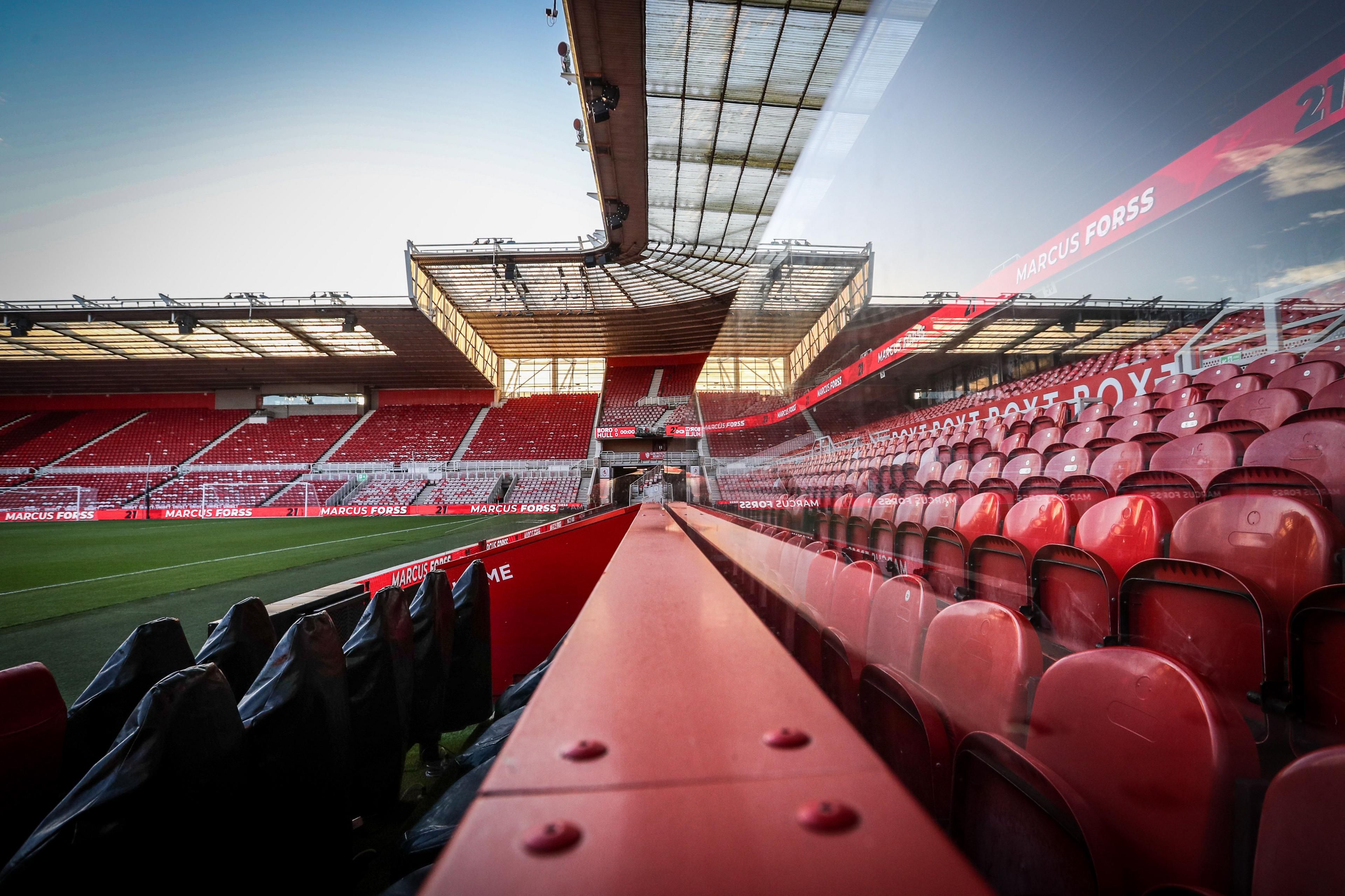 An empty Riverside Stadium. The shot is taken along a steel barrier behind the dug-outs. Rows of red seats and the glass front of a line of executive boxes can be seen on the right. In the distance is the two-tiered stand behind the goal.