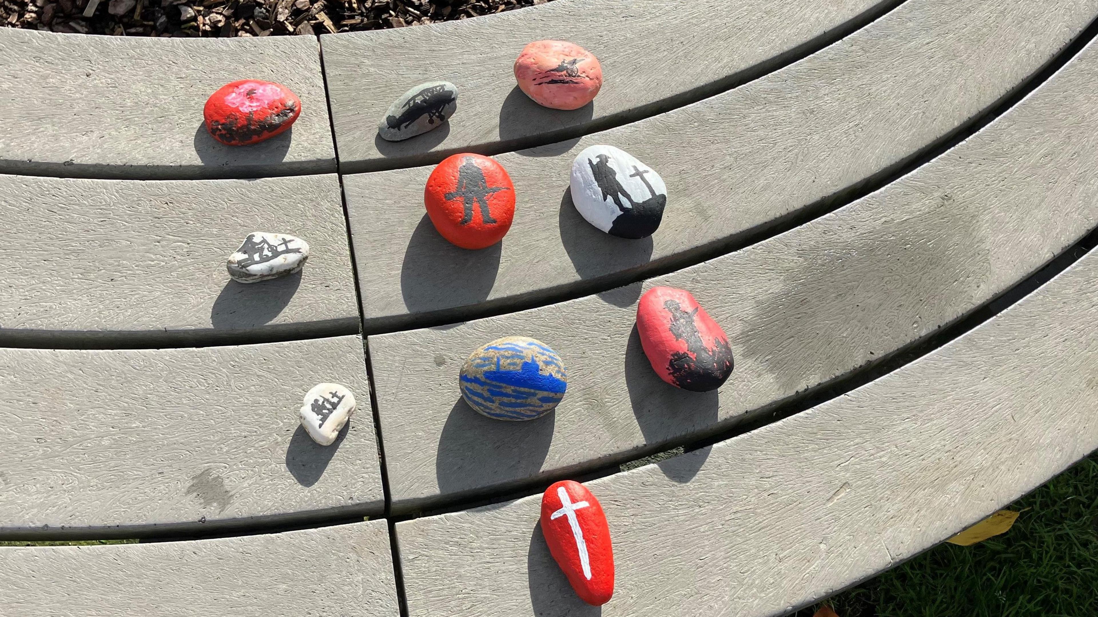 Ten pebbles painted in black and white, blue, red and white, black and pink and a red and pink poppy. Five of the pebbles depict the silhouette of a soldier. One has a warship, another a cross. There is also one depicting an airplane.