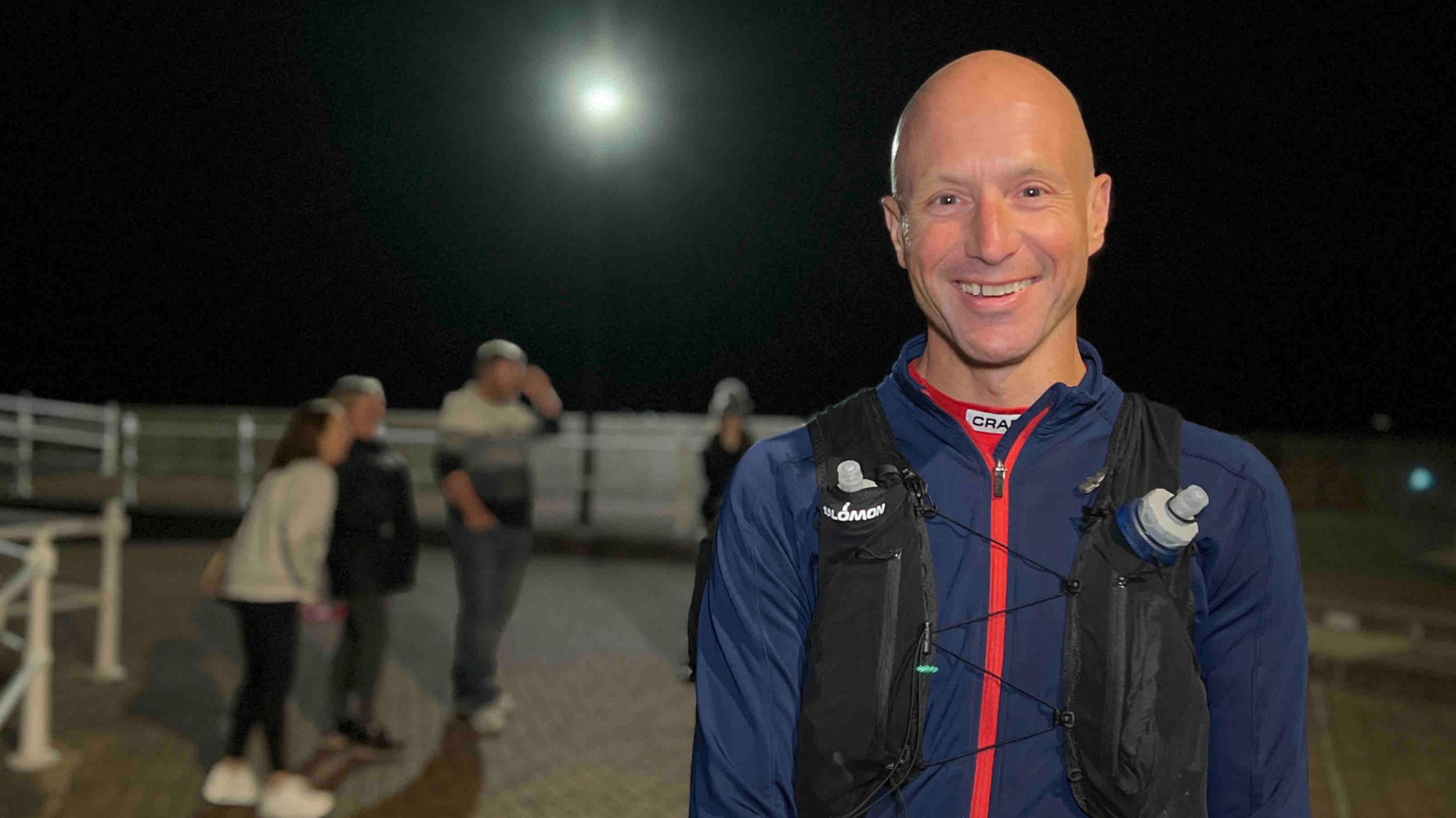 Chris stands outdoors at night wearing a blue jacket with red accents and a hydration vest holding water bottles. He is on a paved area near white railings, with other people visible in the background. The scene is brightly lit from above