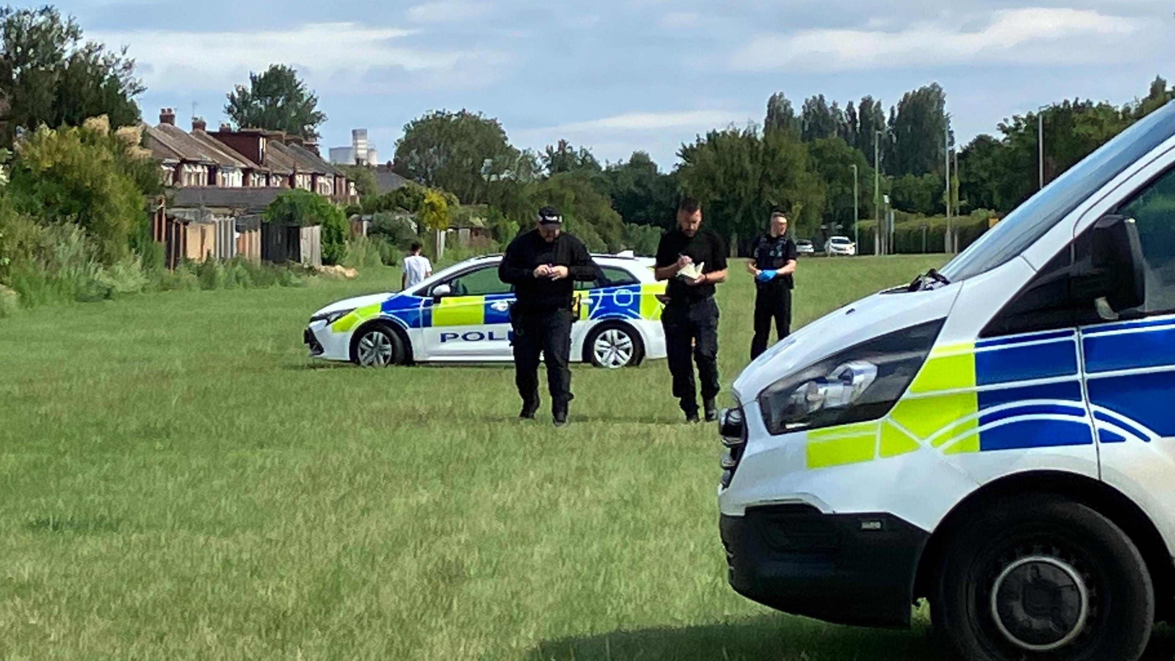 Three police officers dressed in black are walking towards the camera. A police van is parked in the foreground with a police car in the background.