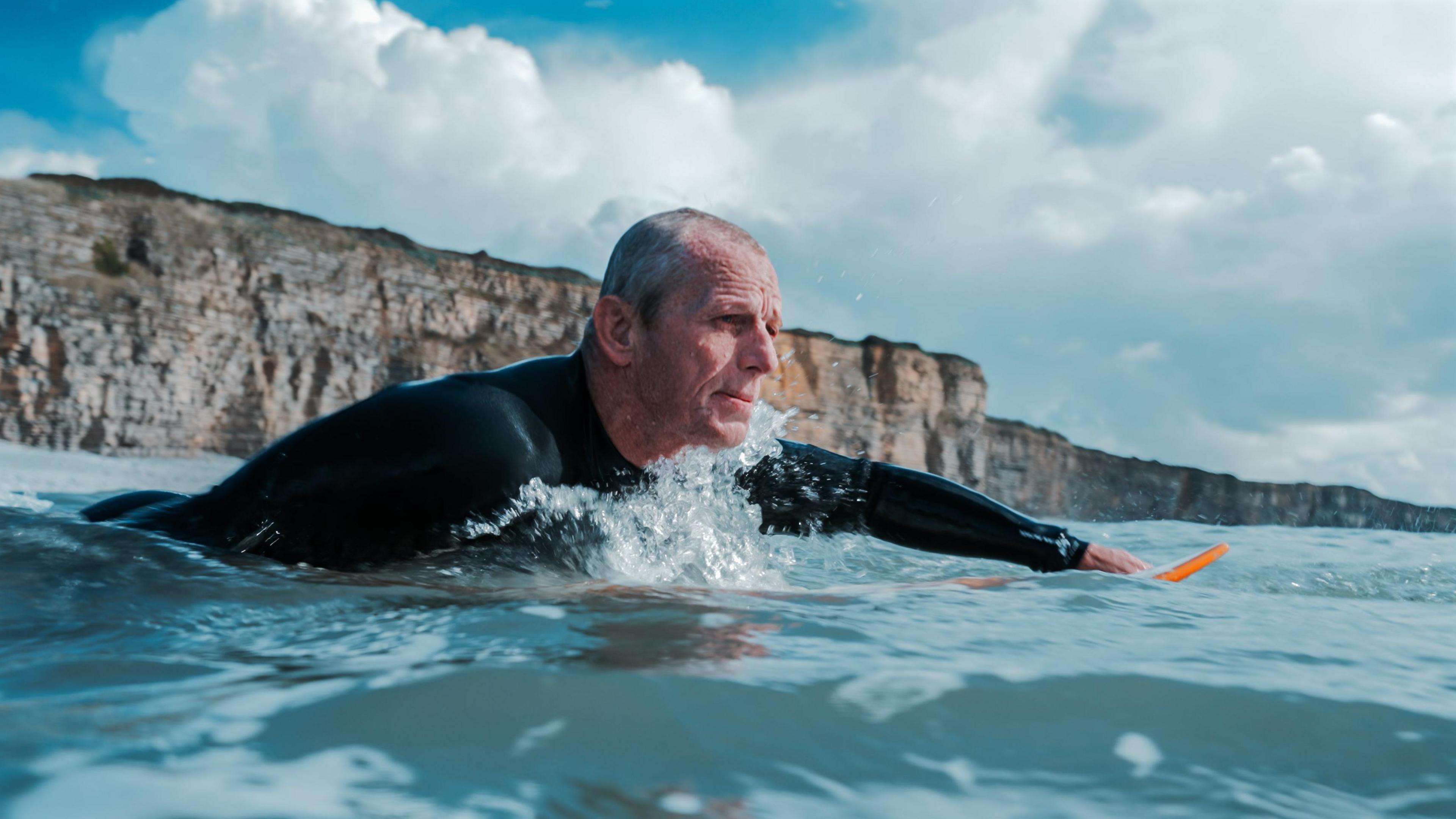 Mark in a wetsuit on a surf board that is mostly under the water. Mark has short grey hair and is paddling out to sea away from a cliff