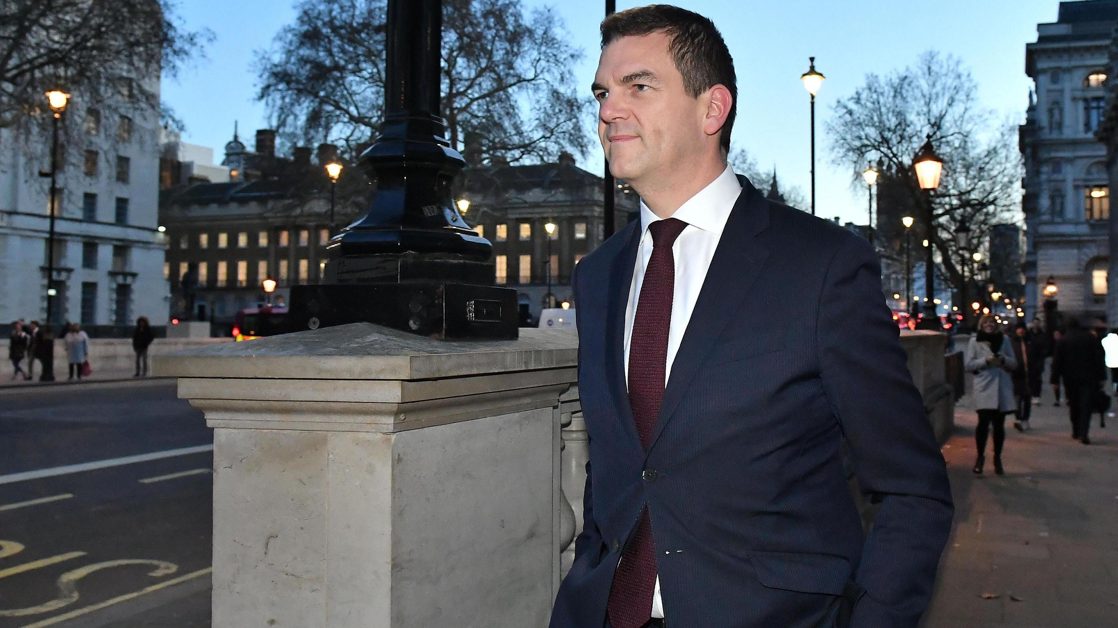 Sir Olly Robbins vesh një kostum blu të errët ndërsa ecën nëpër Whitehall në muzg