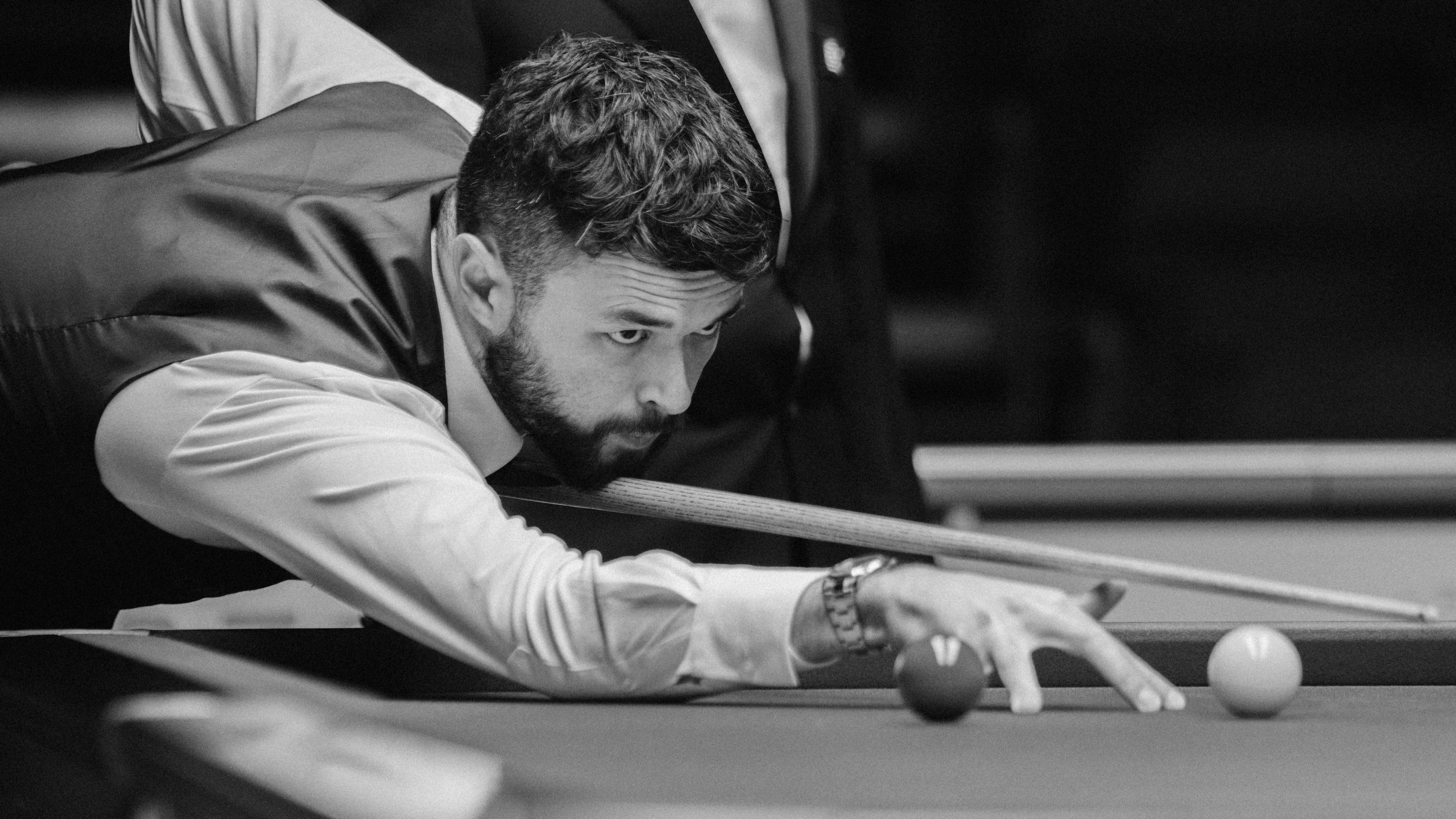 A snooker player, leaning forward towards the right of the frame, lining up a shot. It is in a tournament, he wears a white shirt and black tuxedo. He has a black beard. His expression is focused. The photo is in black and white.