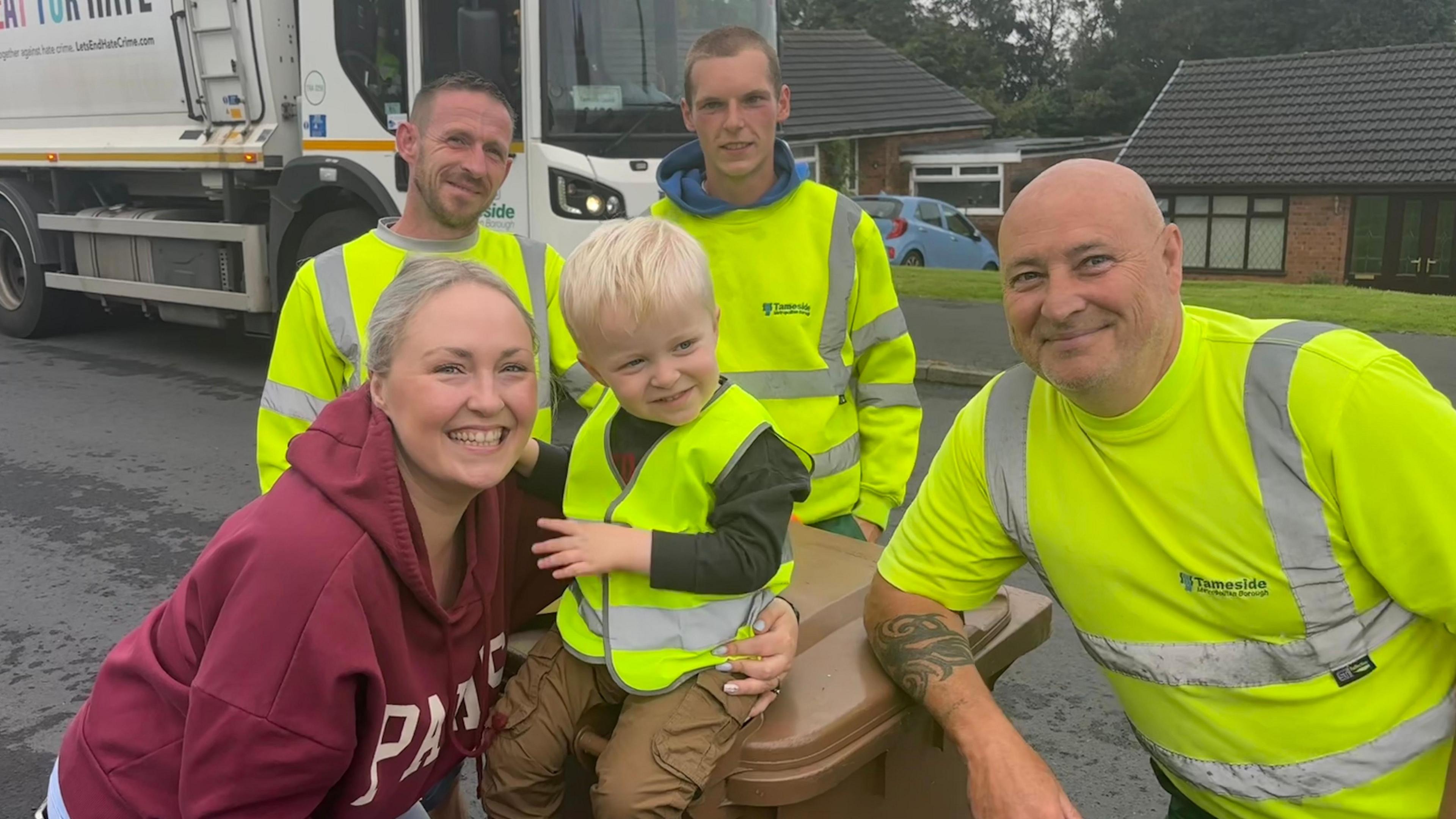Pam, who is wearing a maroon top, with her arms around Teddy, who is sitting on top of a brown bin. They are surrounded by three bin men in the their high-visibility yellow tops. A bin lorry is in the background.