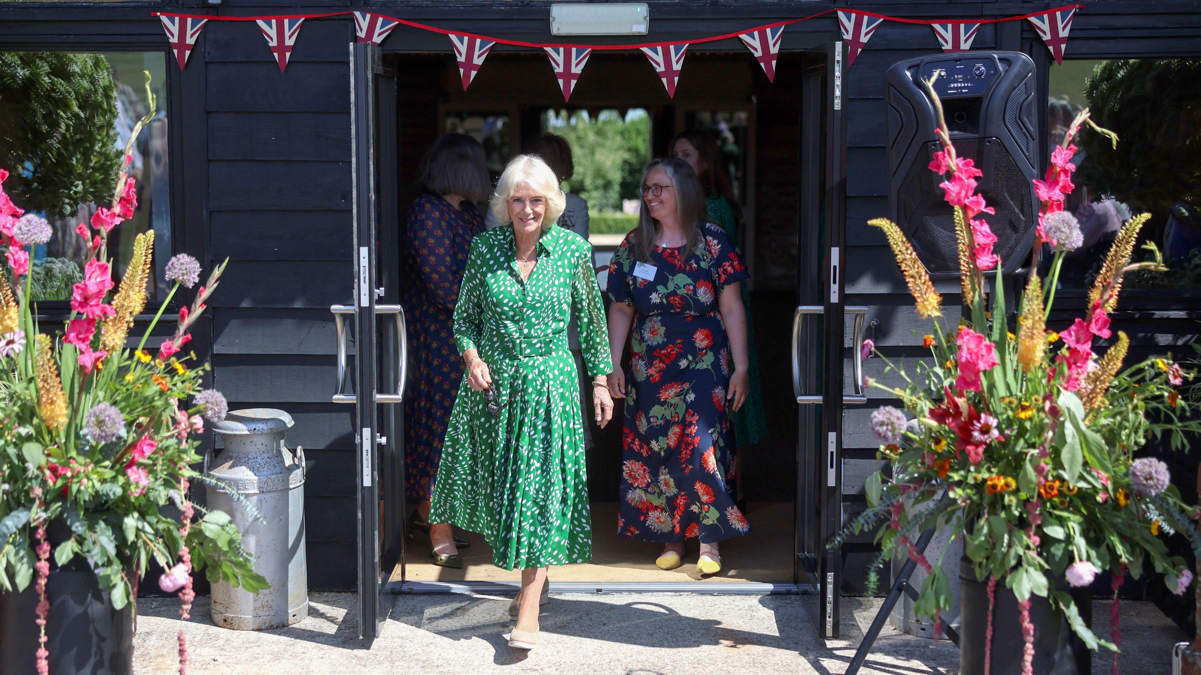 Queen Camilla wearing a green and white patterned long dress and walking outside through double automatic doors. On either side there are large bouquets of flowers, and Union Jack flag bunting above the door.