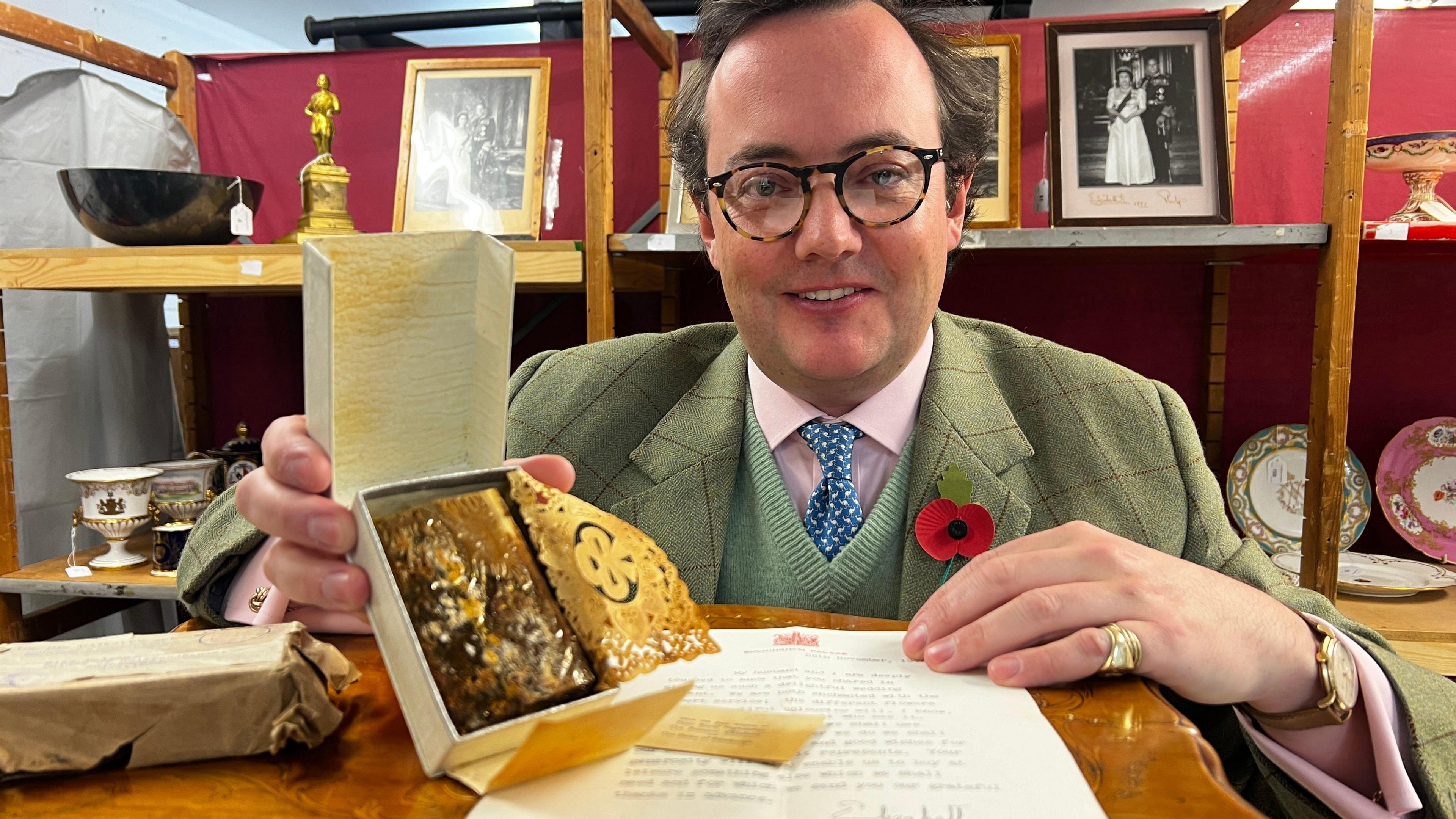 Auctioneer Jonathan Benson holding the cake, which looks mouldy. There is a letter in front of him and he is sat in front of various portraits of Queen Elizabeth II and Prince Philip, as well as china. Mr Benson is wearing a suit with a jumper, pink shirt and tie underneath and a poppy.