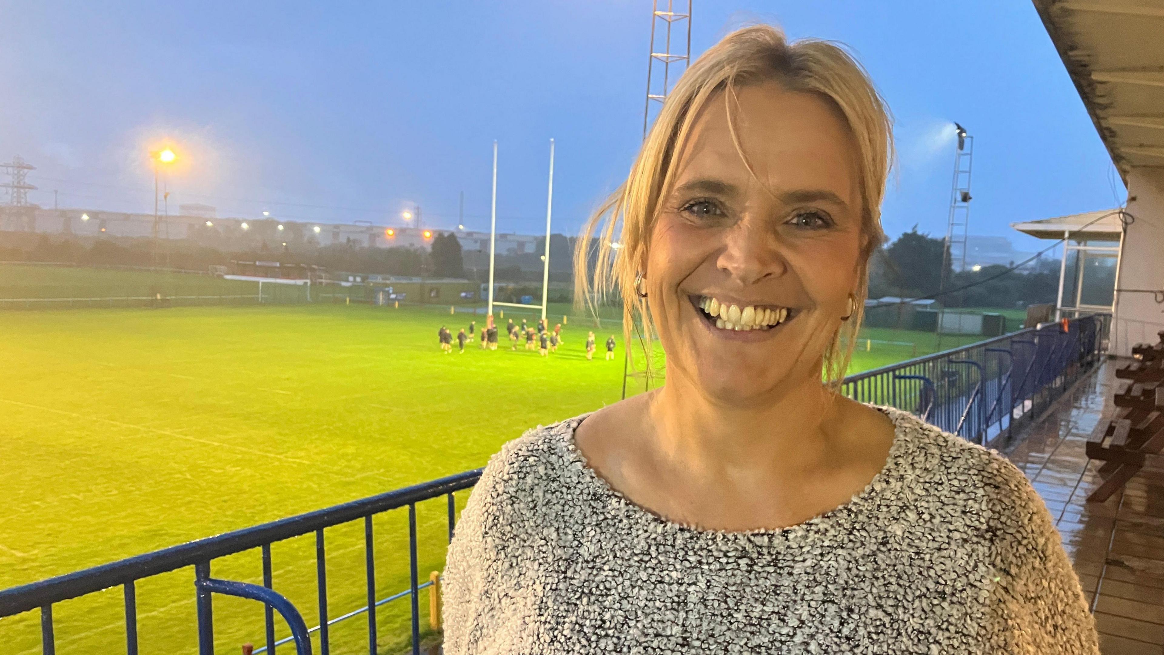 A middle aged woman is smiling with her teeth showing looking at the camera. She's wearing a white and black speckled top and she has blonde hair. She's standing on a large balcony with blue railings which is looking over a rugby pitch. Rugby players training are visible in the background near rugby posts. The sky is darkening and floodlights around the pitch are also visible.