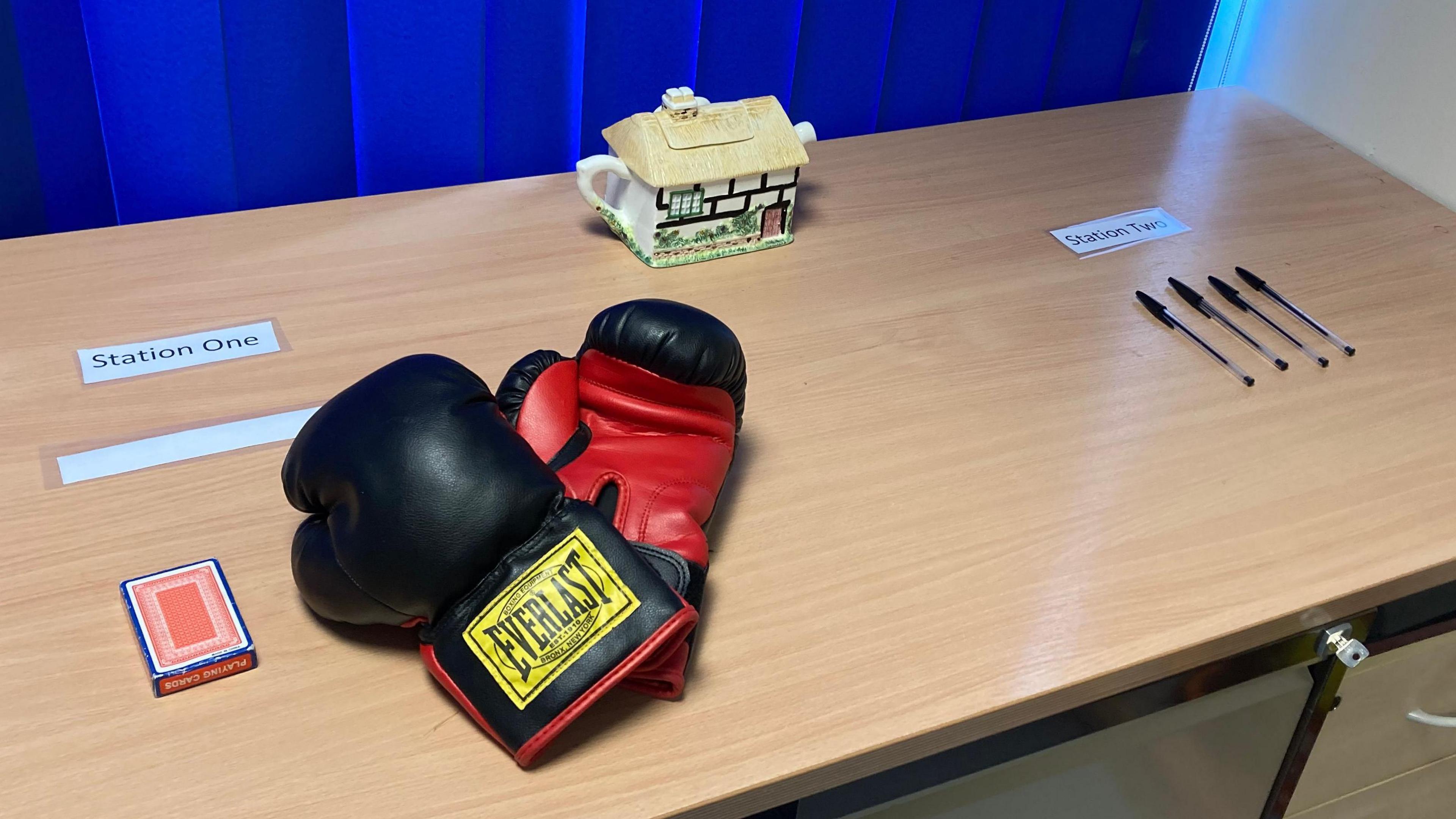 A table is set up with various stations for the escape room. Boxing gloves, a pack of cards, pens and a teapot in the shape of a house sit on the table.