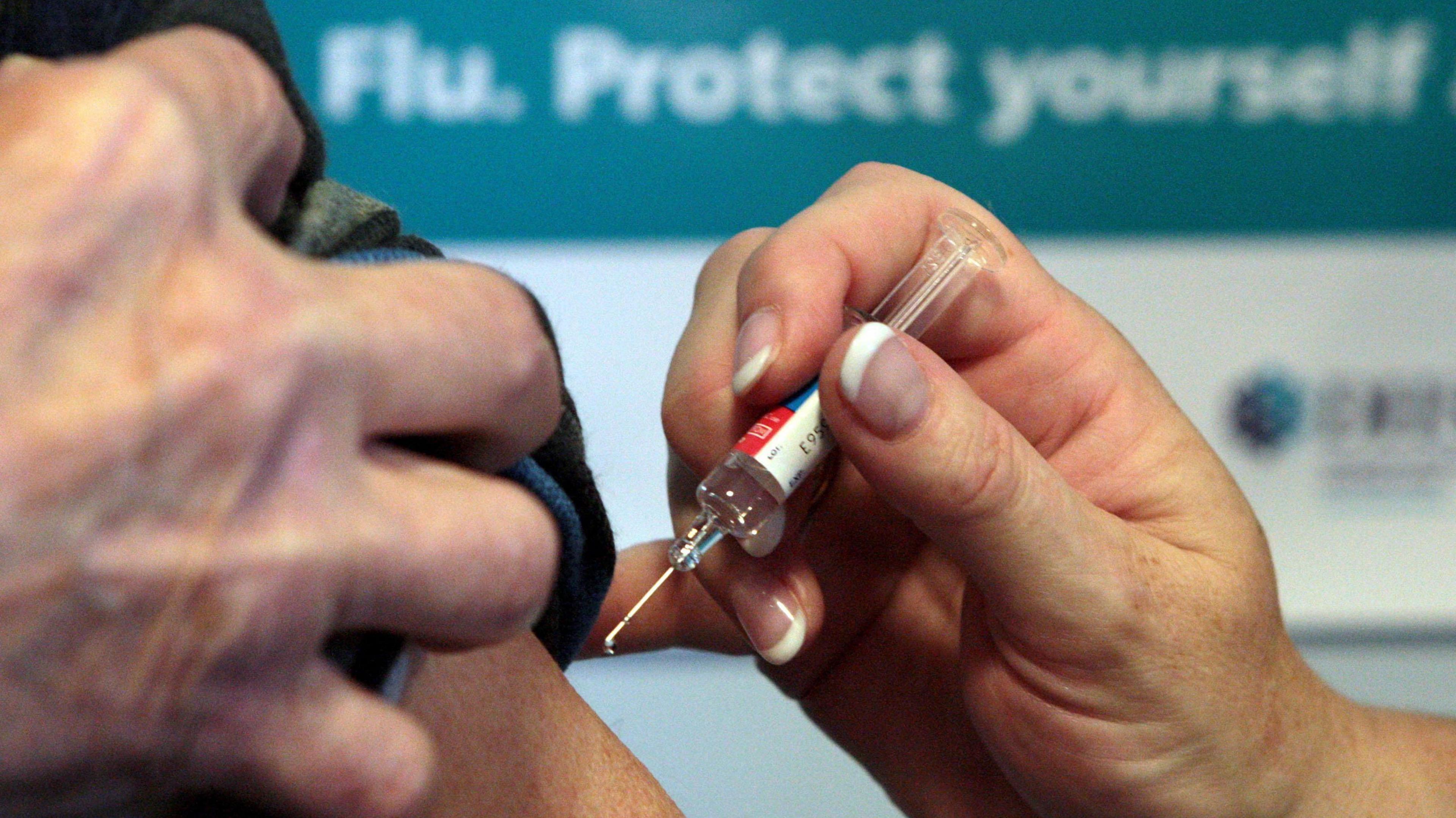 Close up of a man rolling up his sleeve, while another pushes a small needle into the top of his arm. A blurry banner behind reads: Flu. Protect yourself.