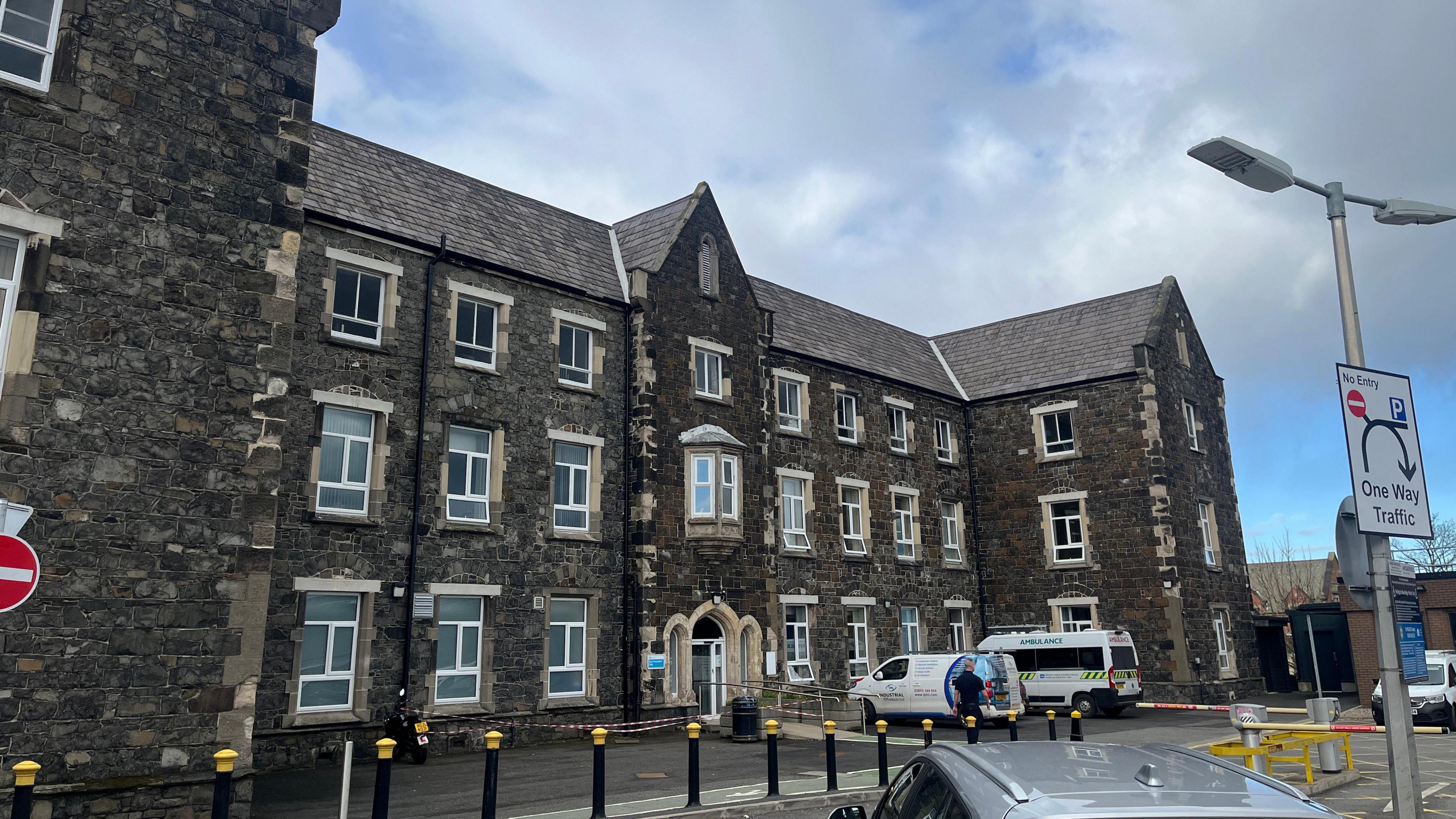 Fever Hospital building on the Lisburn Road. It is a three-storey dark stone building. There is a white van and a white ambulance parked outside its front doors.