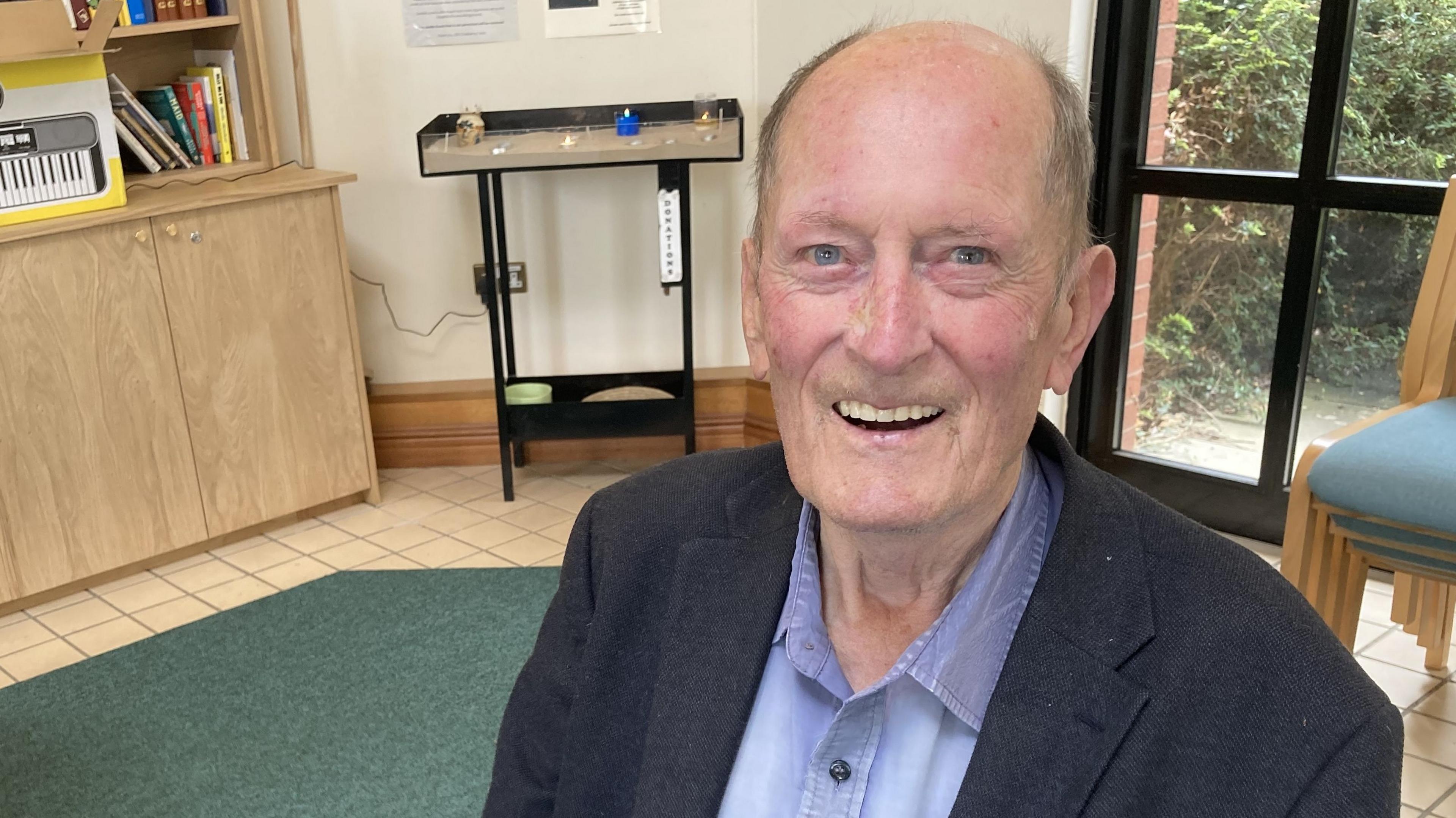 Tim Edwards is sitting on a chair in the hospital chaplaincy and is smiling. He has a near bald hair with light coloured hair around the edges and wears a blue shirt with a dark jacket.