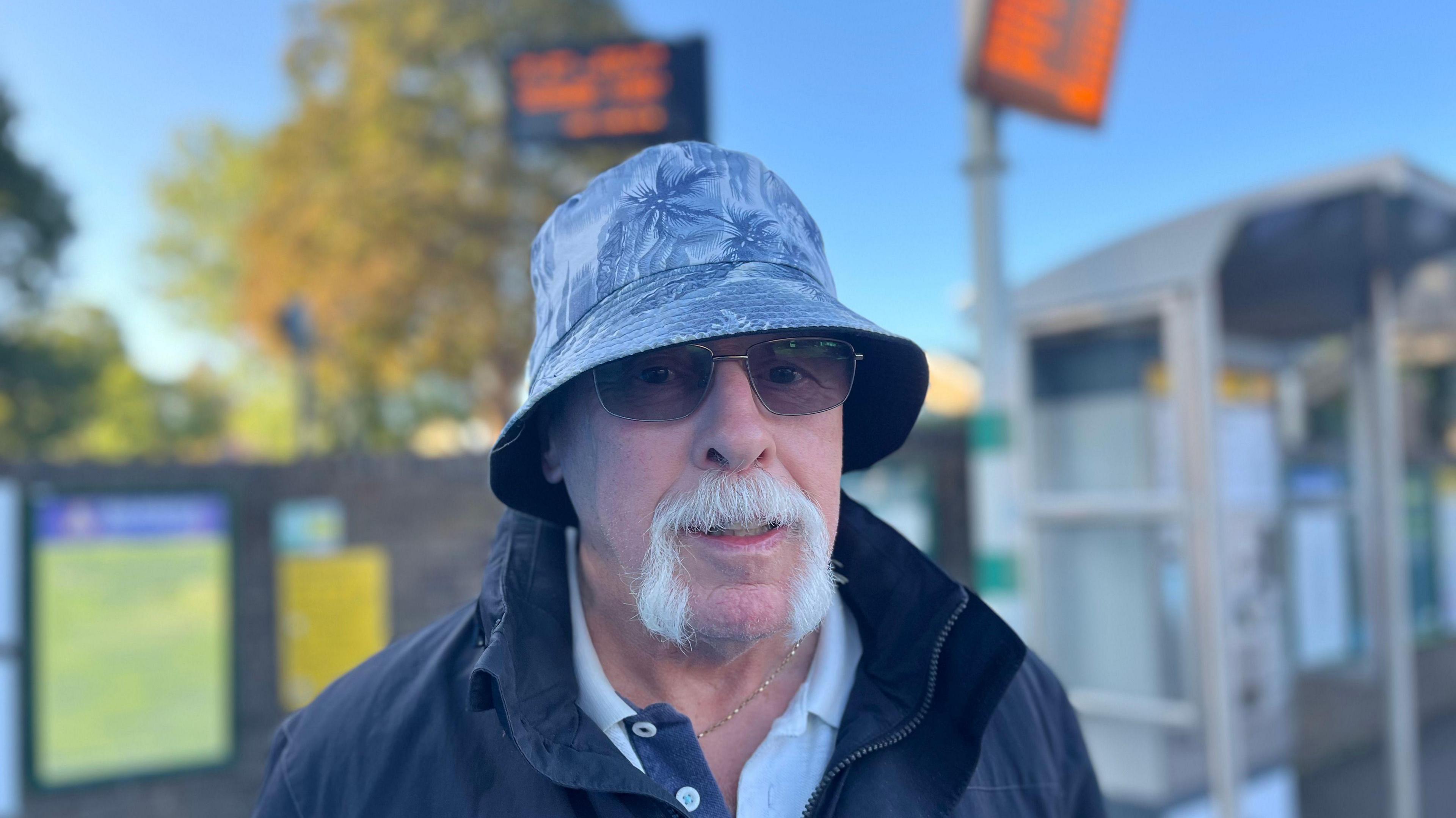 Mike Chiapporiwearing a bucket cap and bifocal glasses. He has a grey goatee and is wearing a coat and standing in front of Ifield railway station with electronic orange boards behind him