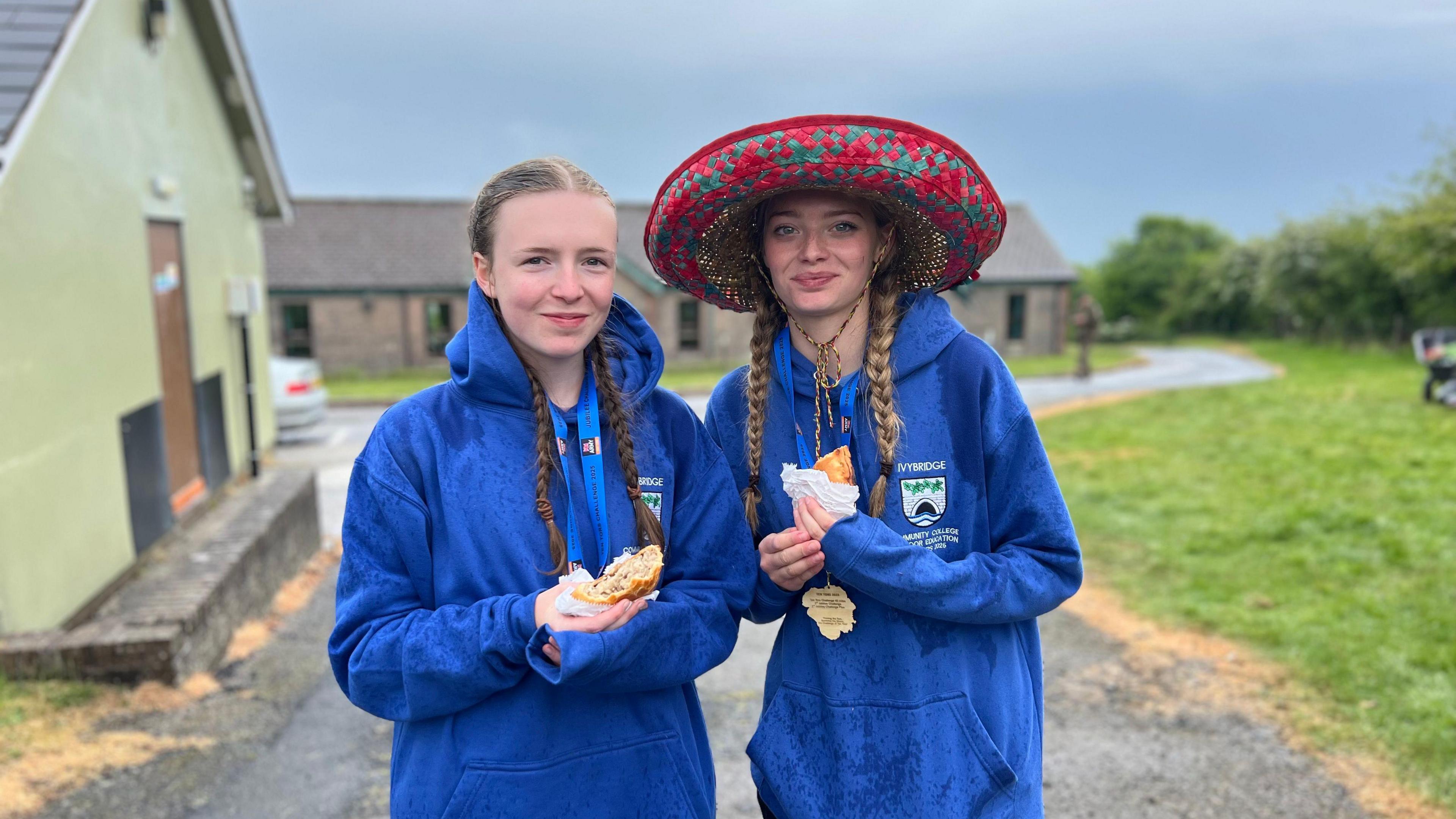 Myrtle and Milly wearing blue jumpers with food in their hands. Milly on the right is wearing a large hat.