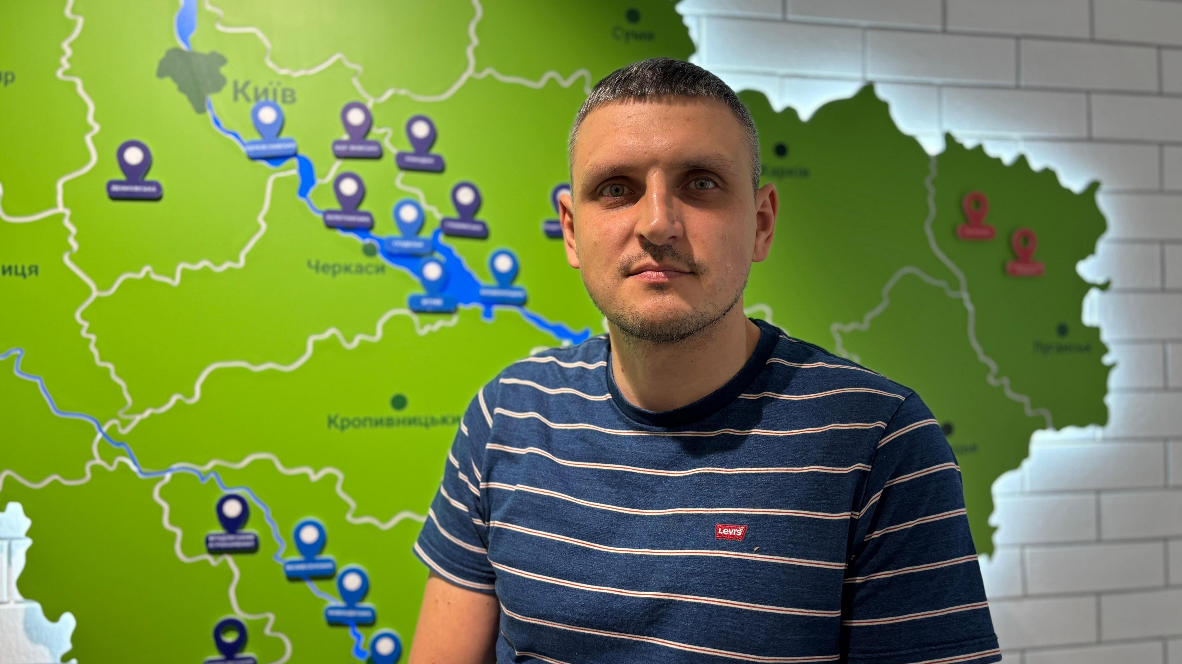 A man in a striped blue and white t-shirt stands in front of a map of Ukraine, which is lit up on the wall behind him.