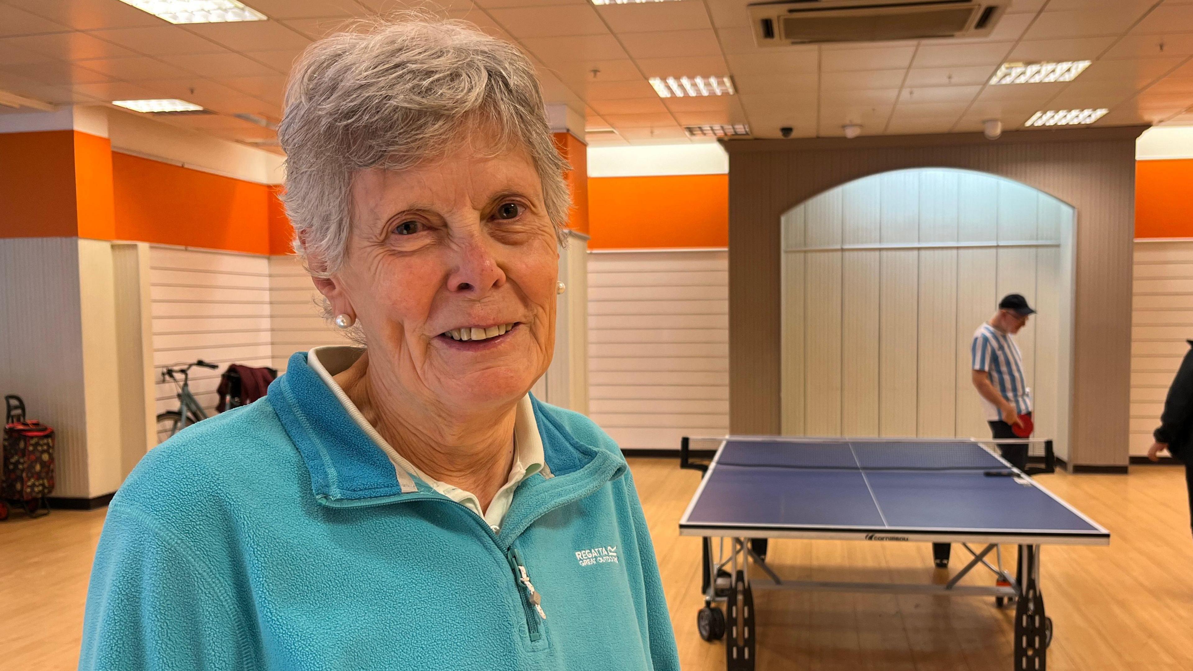 Valerie Lawrence is smiling at the camera and standing in an open space with a table tennis table in the background and a wooden floor. She has short, grey hair and is wearing a blue top