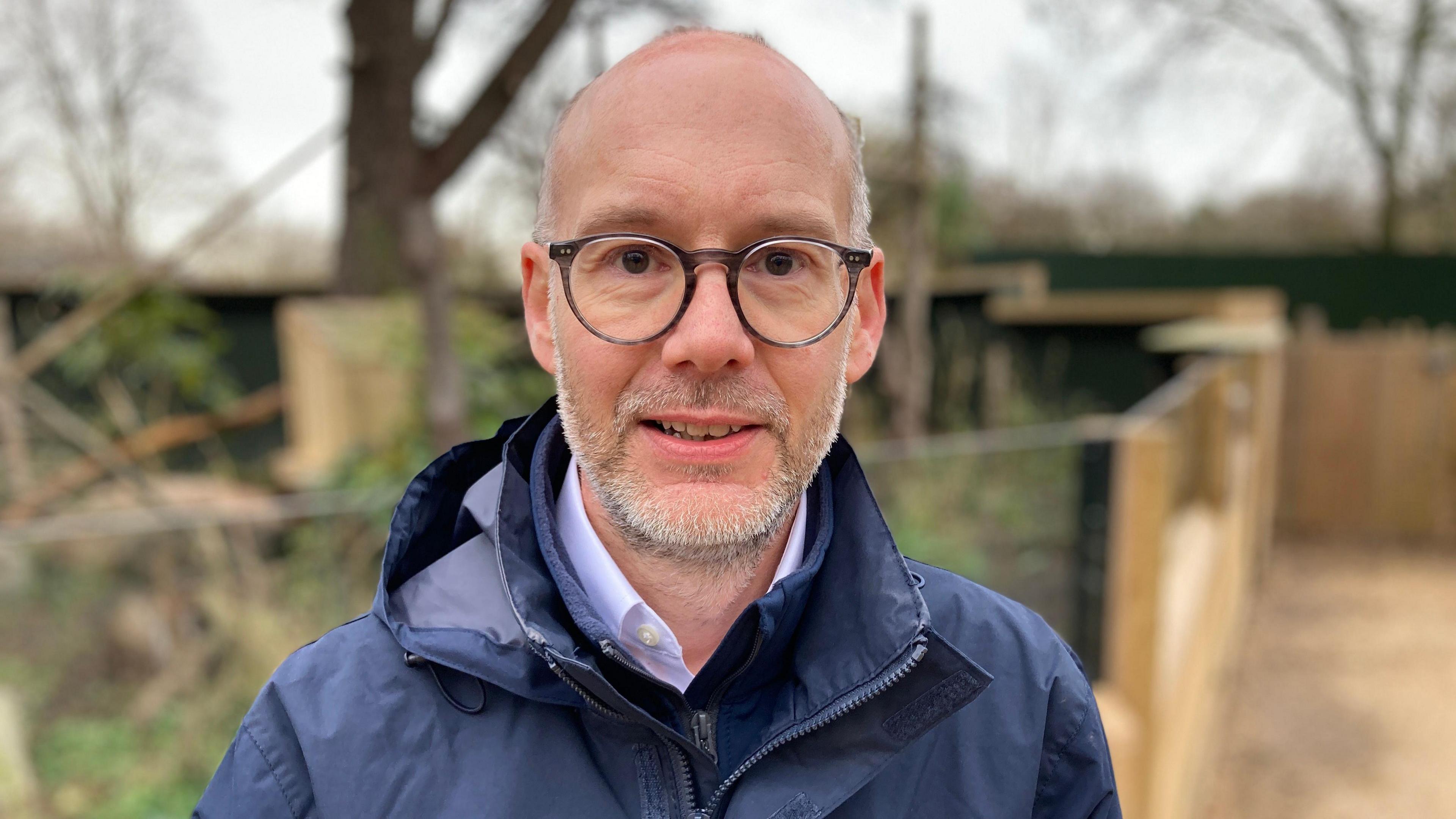 Justin Morris wears a navy blue raincoat over a white shirt. He has short grey hair and black round rimmed glasses.