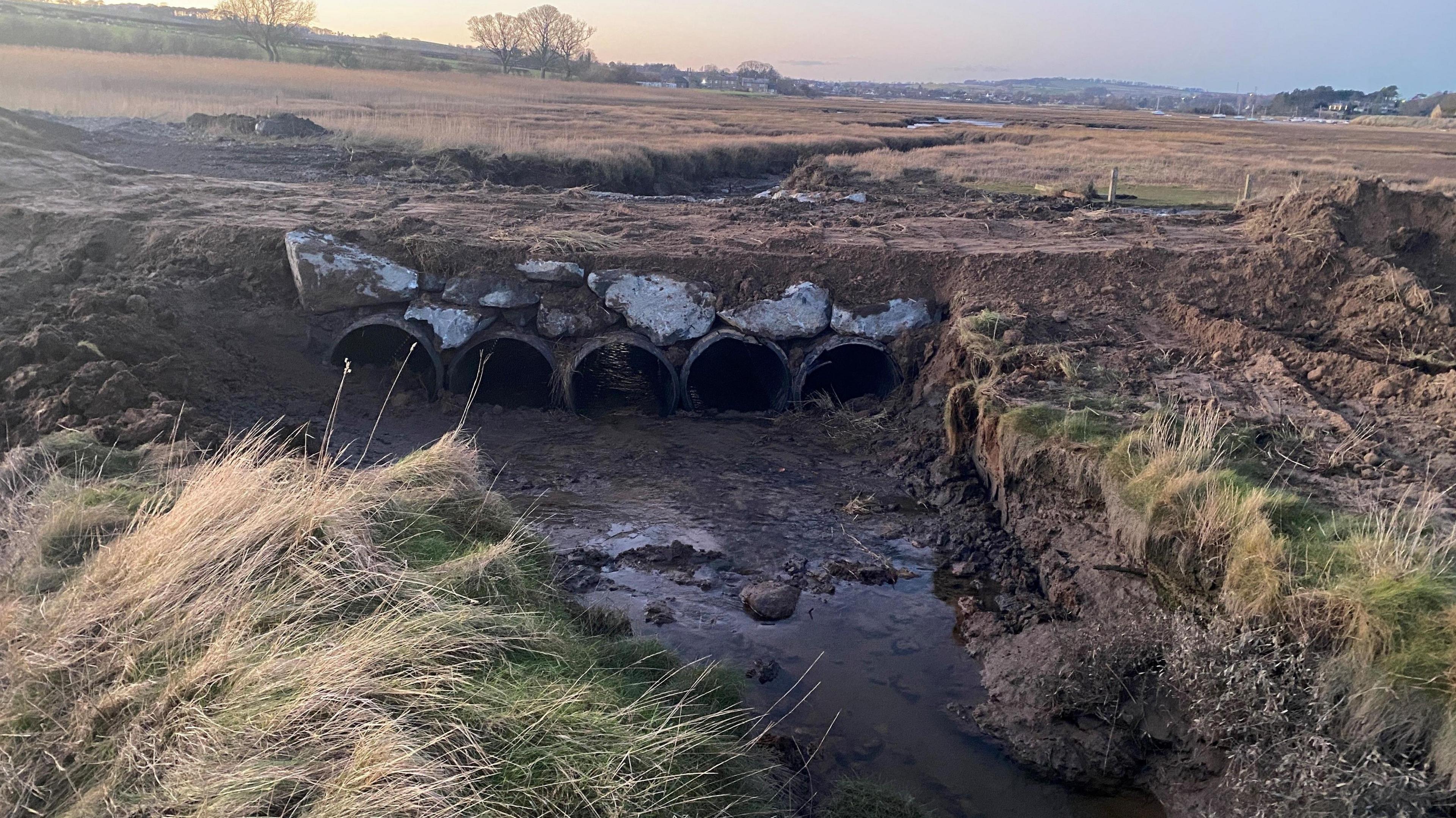 Five newly installed large concrete pipes sit under a footpath which passes over some tidal mudflats. The construction allows water to pass under the raised path.