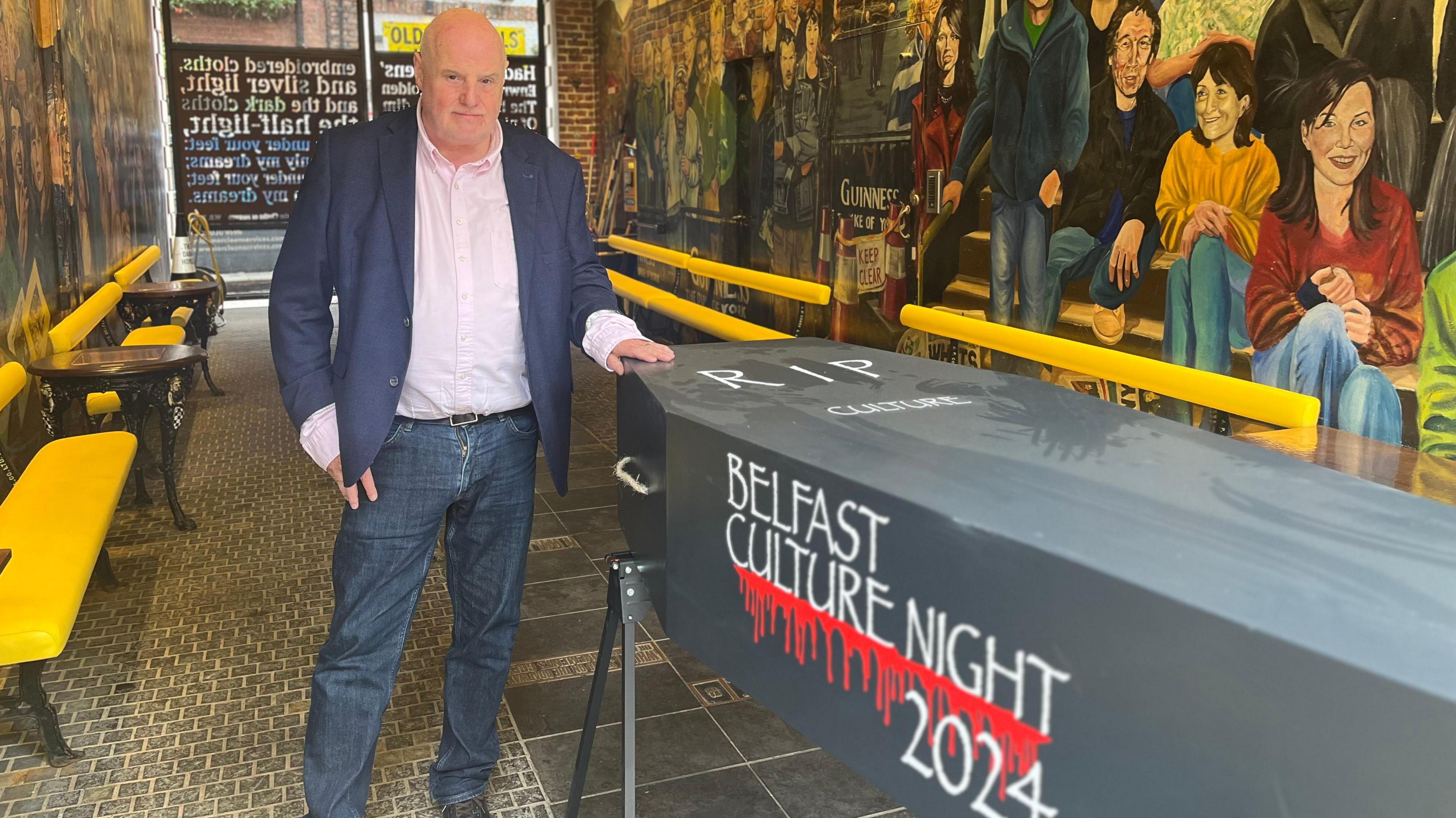 Willie Jack wearing denim jeans, a navy blazer and a pink shirt, he is standing next to a black coffin that says "RIP Culture" and "Belfast Culture Night 2024" with blood dripping from it