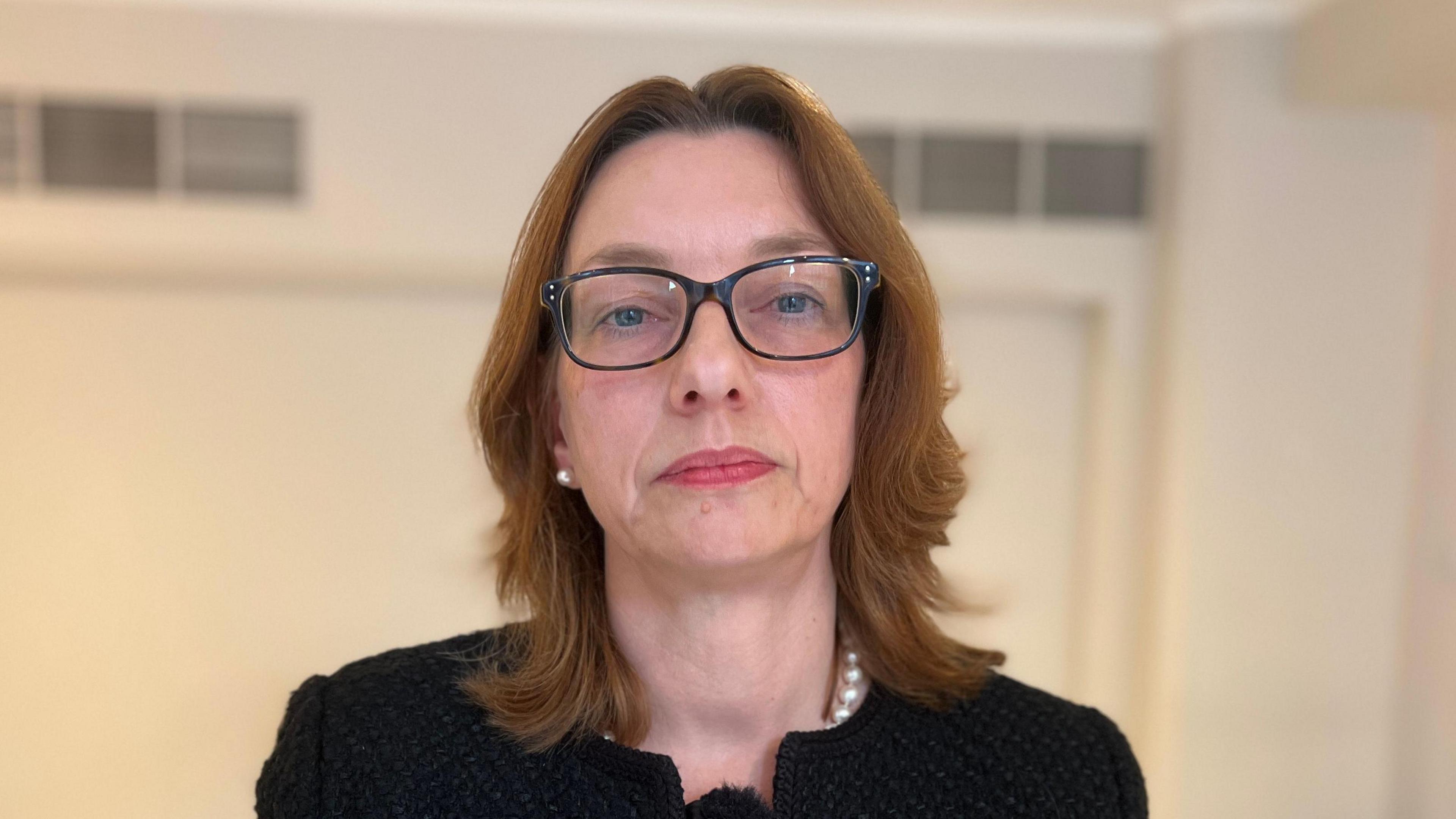 Jackie Yates from the shoulders up - she is wearing a black jacket, glasses and has medium length brown hair. A cream-coloured wall is out of focus in the background