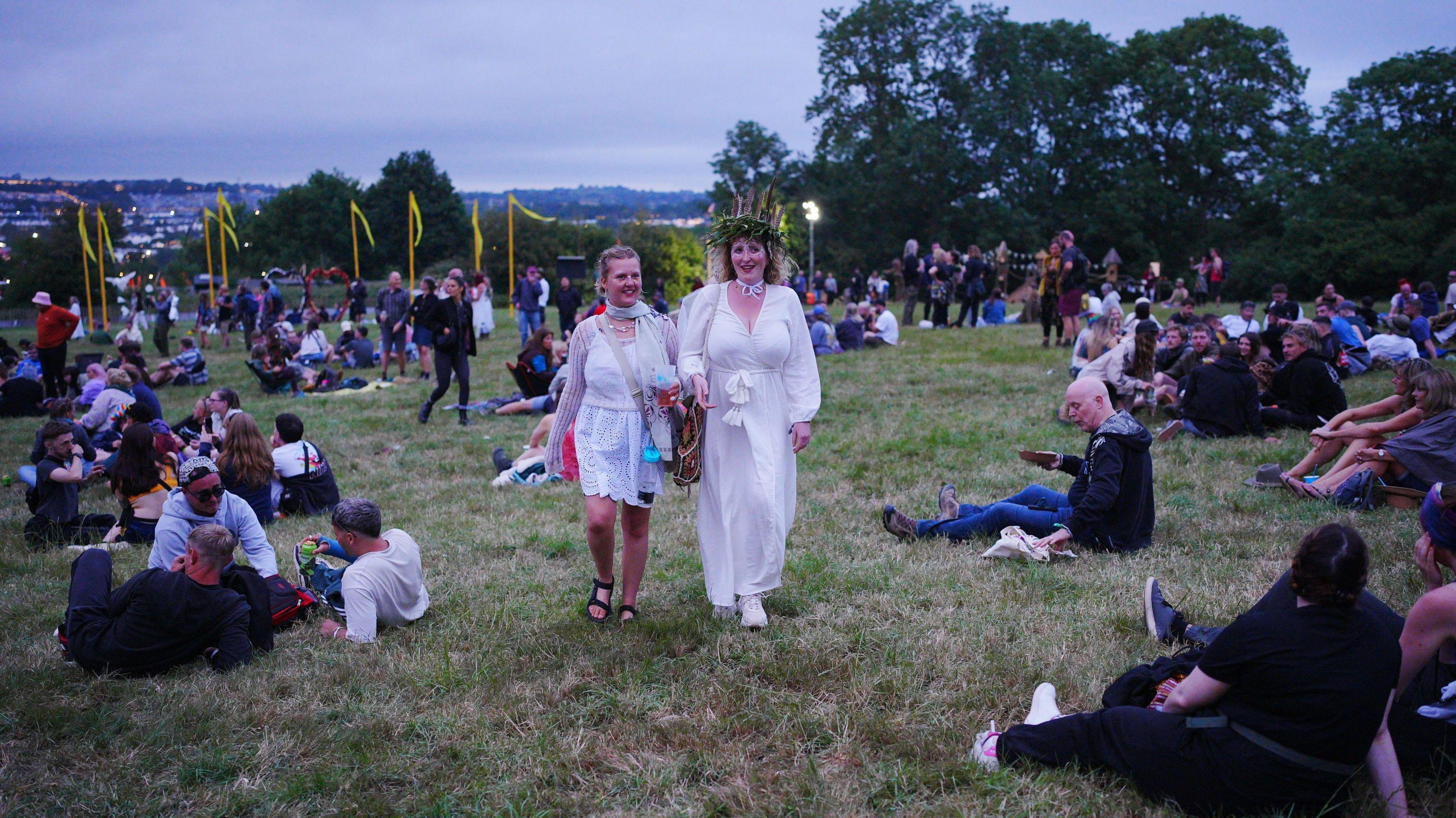 Revellers at the Stone Circle enjoy Glastonbury Festival. Two women are walking towards to the camera. They are dressed in white flowing dresses. 